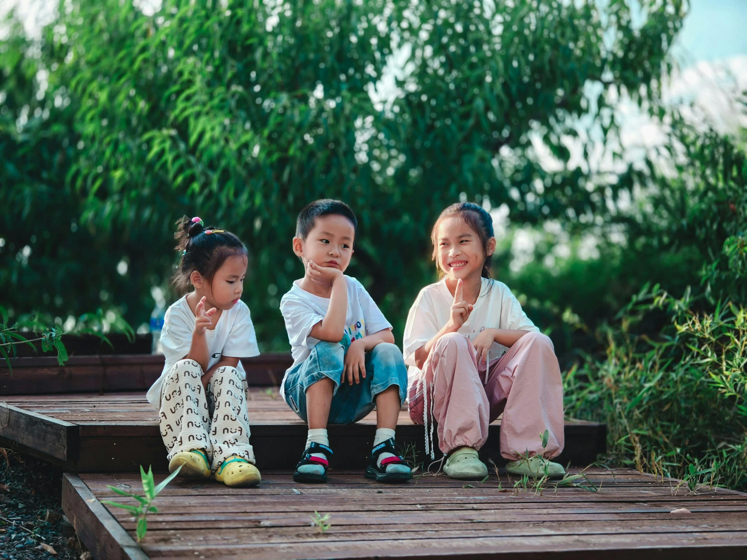 Three young children sit on a wooden step. The background is full of greenery. They are all looking in different directions. Two look bored and one is smiling brightly.