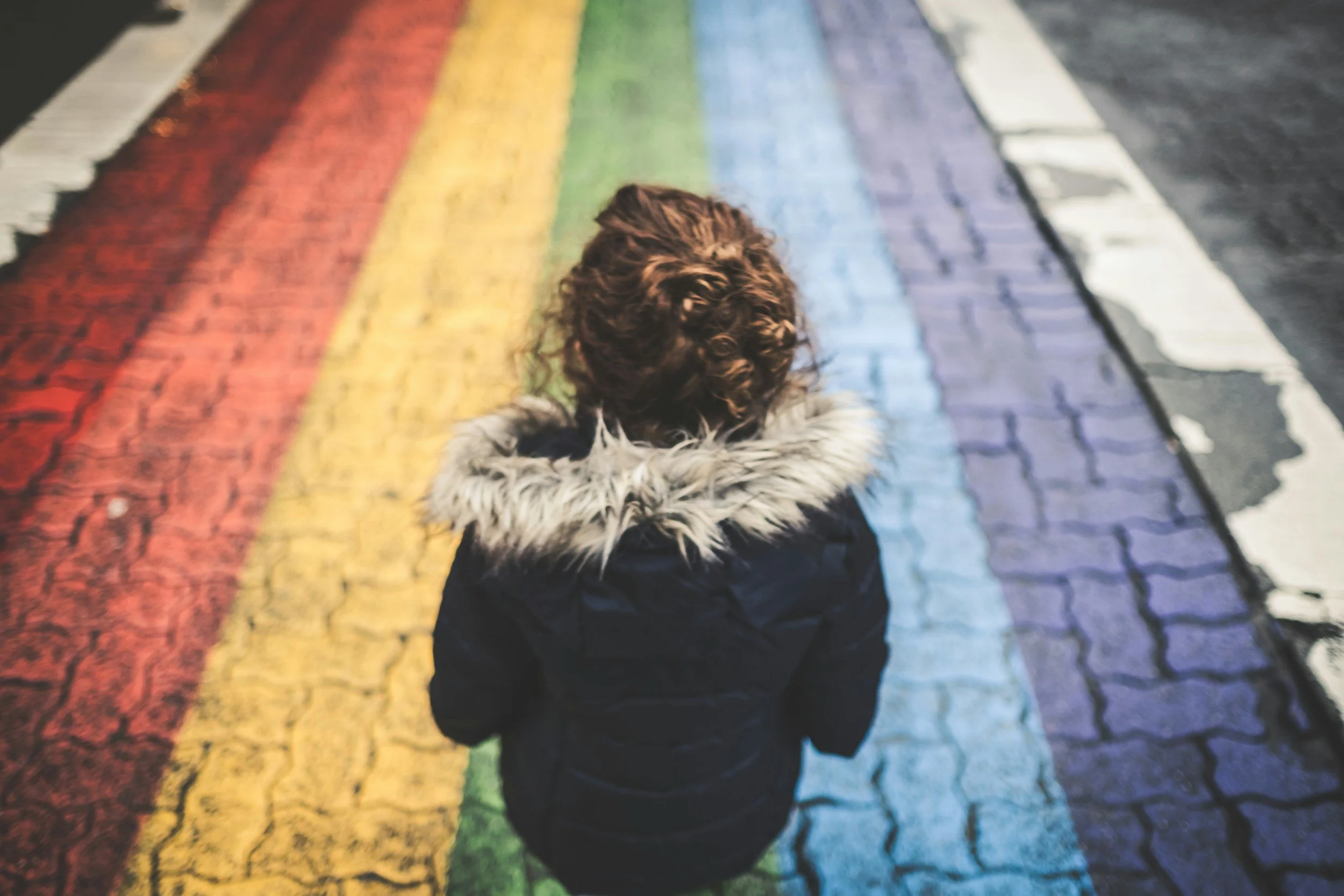 A child stands on a sidewalk that has rainbow strips. They are facing away from the camera. The sidewalk is made of bricks. The child is wearing a winter coat with a fur hood. Their hair is brown and pulled up in a messy bun.