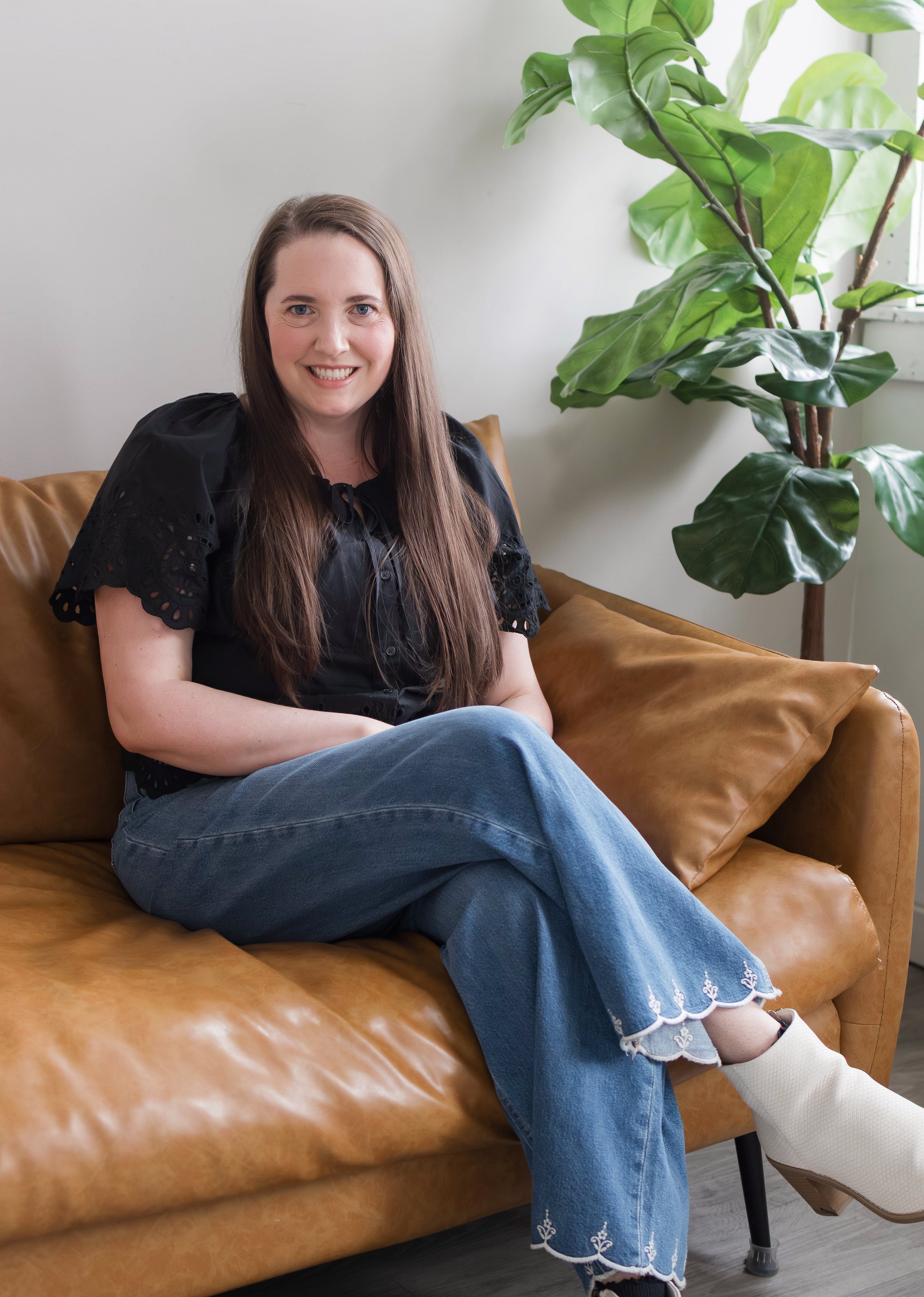 Therapist photo, therapist sits on couch, she is wearing jeans and a black shirt, her legs are crossed, she is smiling and looking at the camera