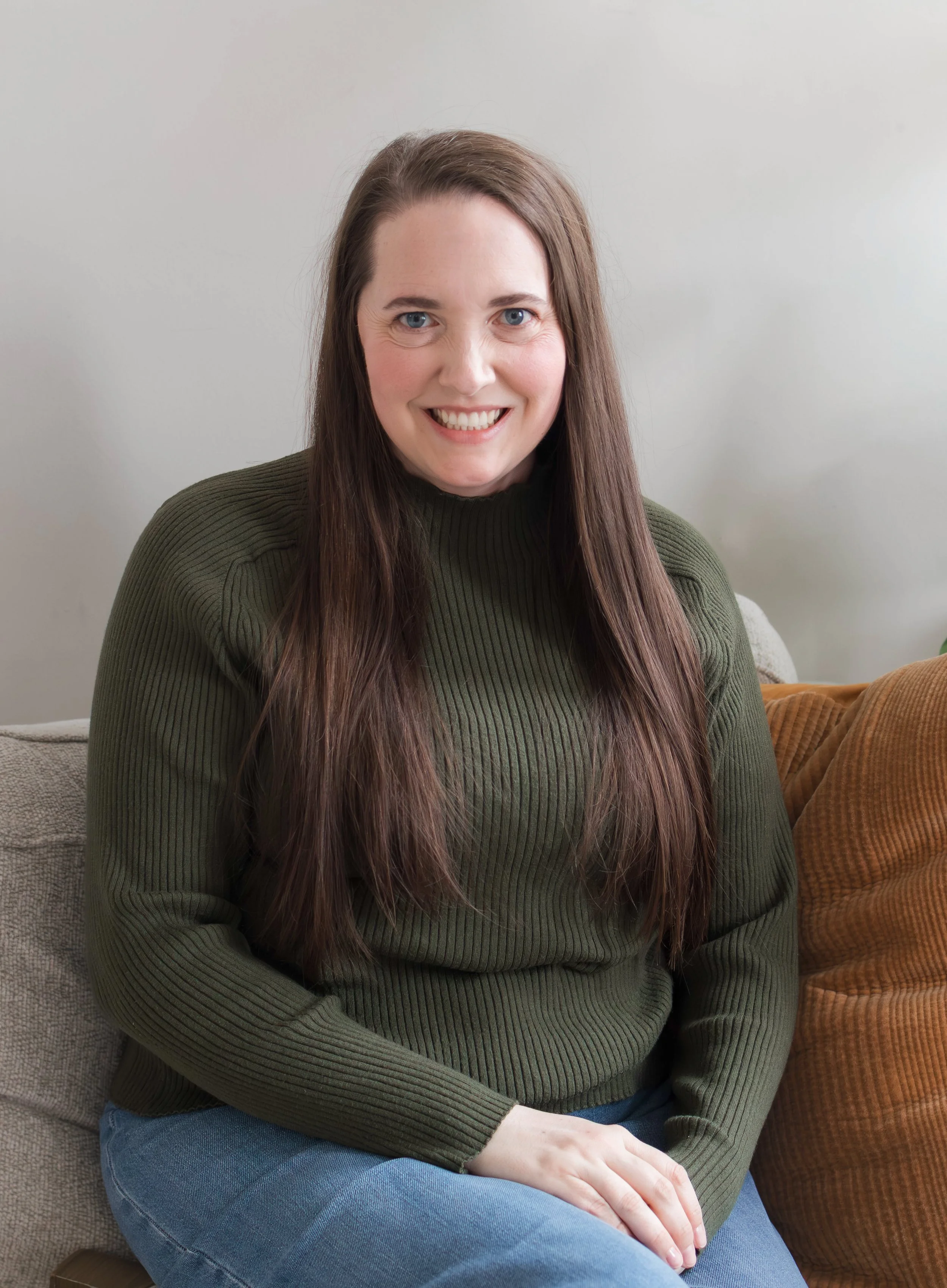 Therapist headshot. Therapist is wearing a green sweater and sitting on a gray couch with a brown pillow. She is looking at the camera and smiling