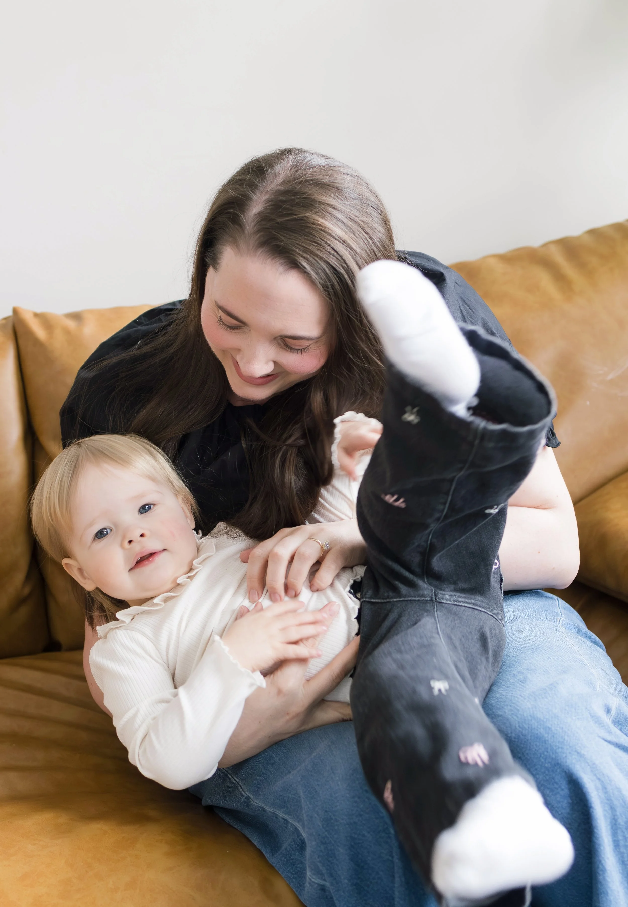 Woman and young girl playing together on a brown leather couch, the woman is smiling and leaning over the girl, who is lying on the couch with her head on the woman's lap, wearing a white long sleeve shirt and blue jeans.