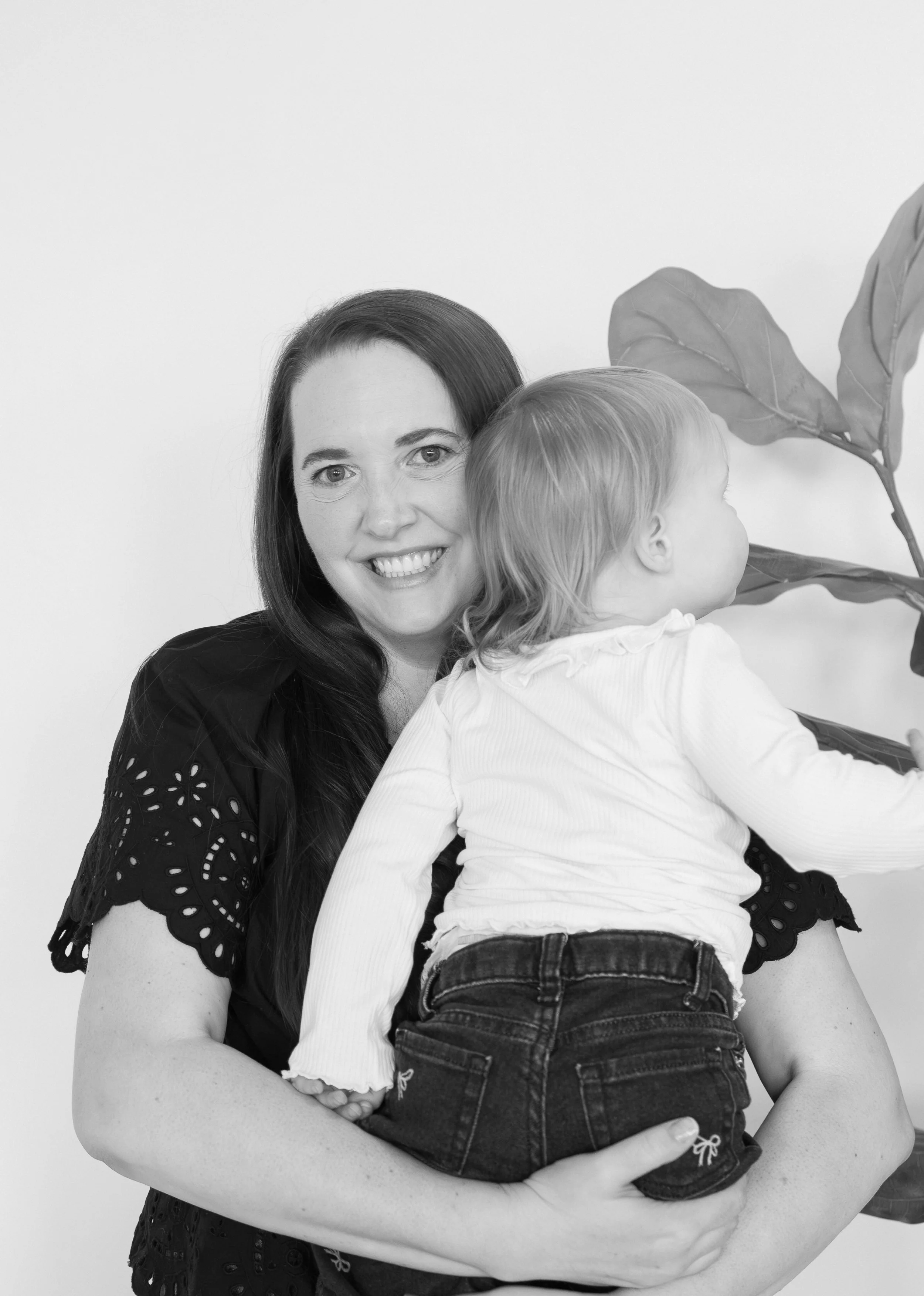 Therapist holds her daughter over her shoulder, she is smiling at the camera. The photo is black and white, a plant is in the background