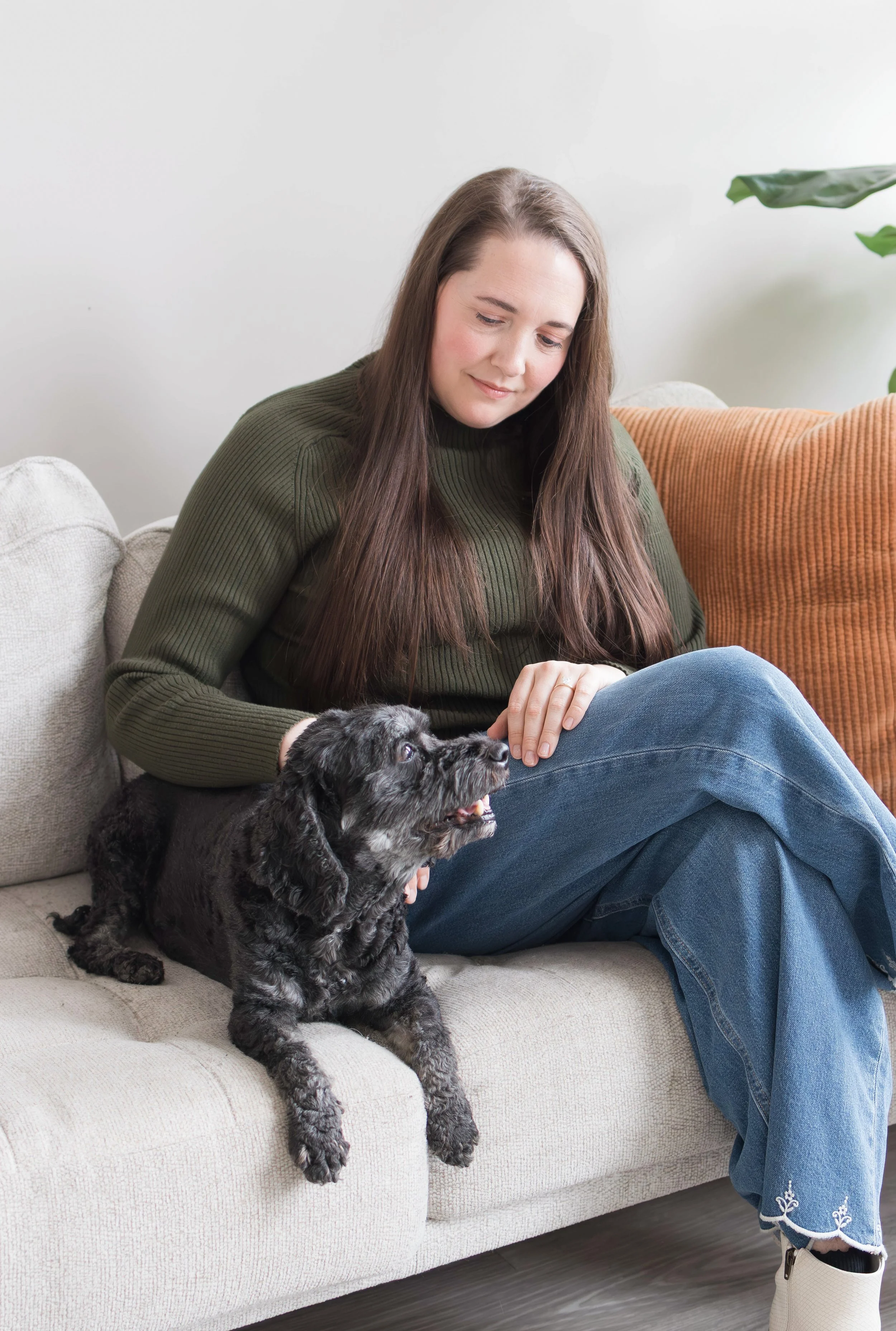 A woman with long brown hair, wearing a green sweater and blue jeans, sitting on a beige couch, petting a black and gray puppy.