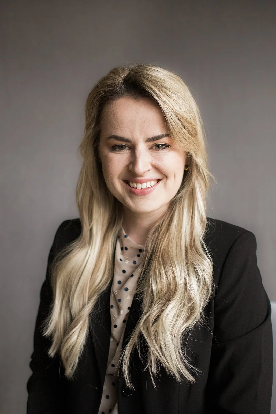Provider headshot, psychiatrist looks at the camera smiling, the background is a muted gray color, she has blond hair , a black jacket, and is smiling at the camera