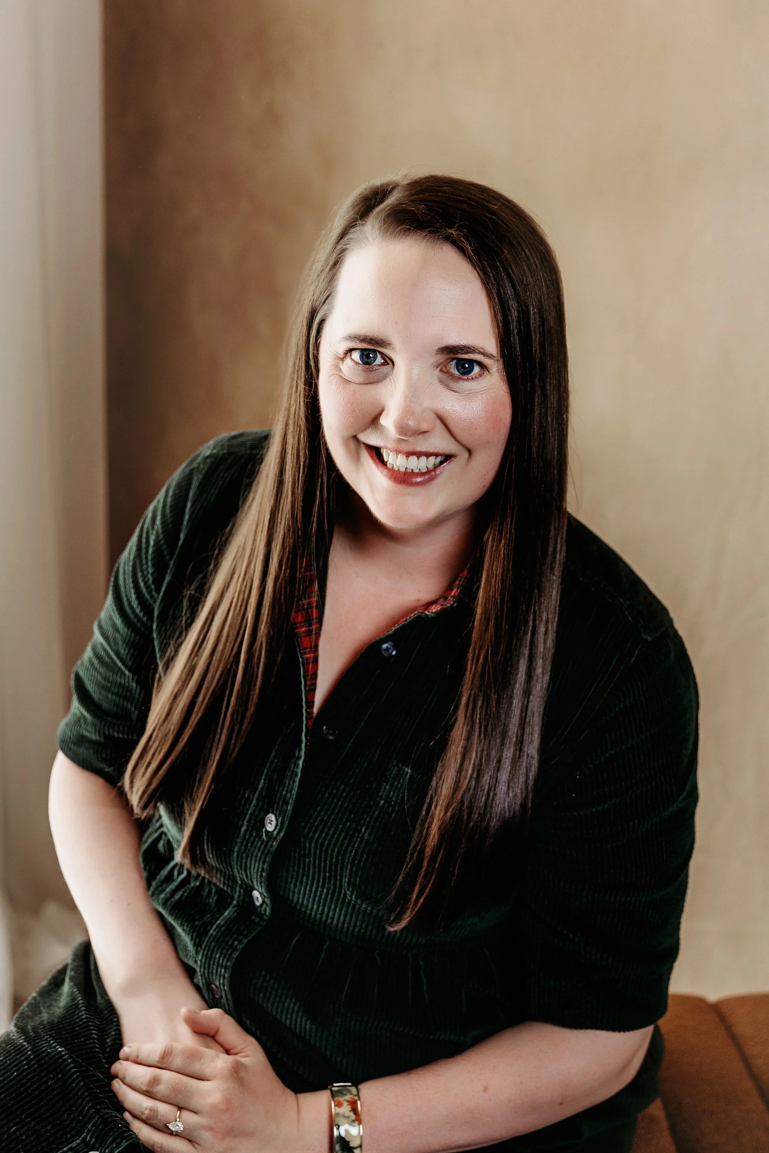 A woman with long brown hair, blue eyes, and a friendly smile sitting indoors. She is wearing a dark green, velvety jacket over a plaid shirt. She has a ring on her left hand and a bracelet on her right wrist, with a neutral-colored wall in the background.