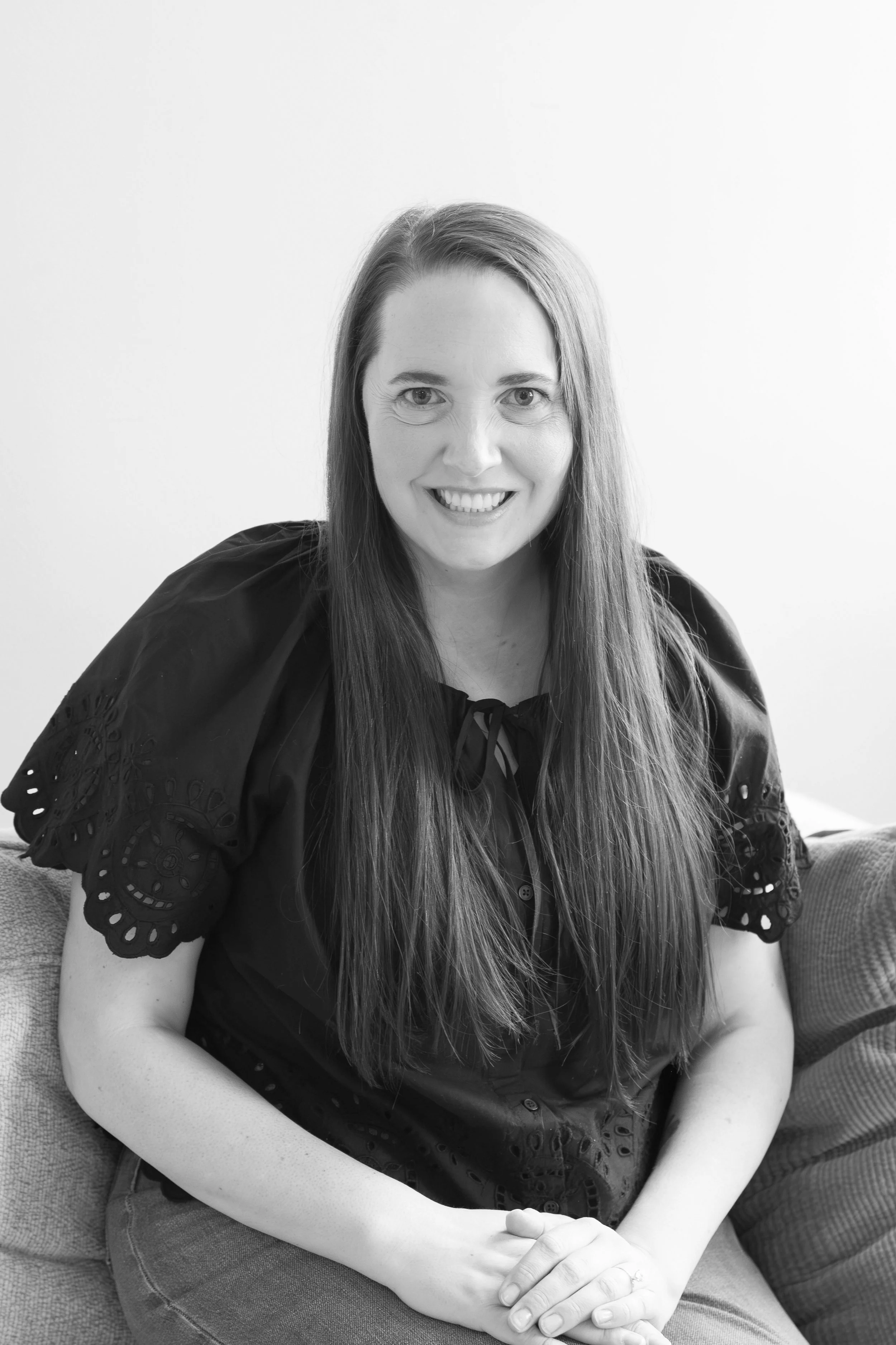 Therapist black and white headshot. Therapist is wearing a black shirt and staring at the camera smiling. She is sitting on a couch, her hands are in her lap, she has long brown hair and she is smiling