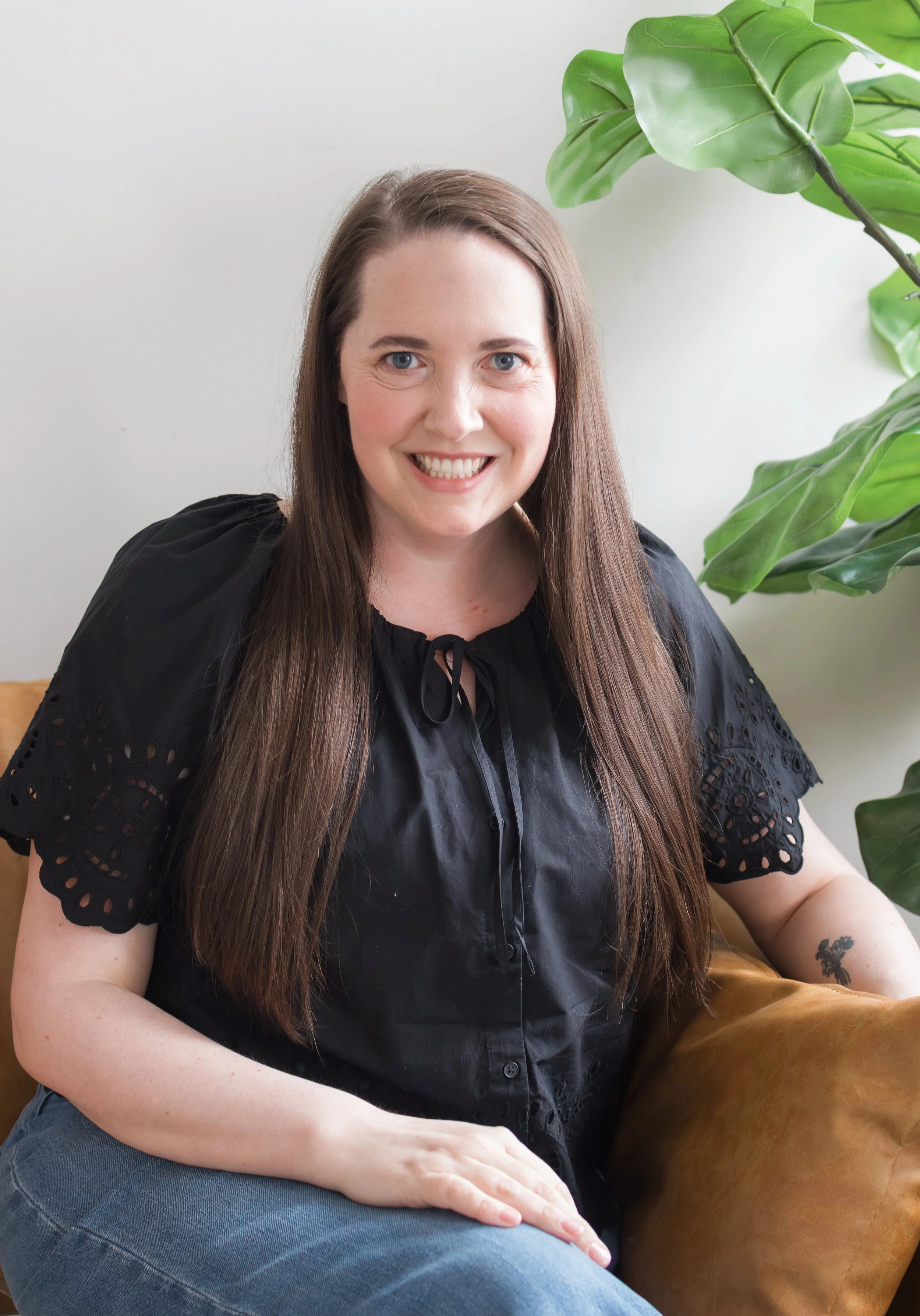 Therapist head shot, looking at the camera and smiling, sitting on a brown couch with a plant in the distance