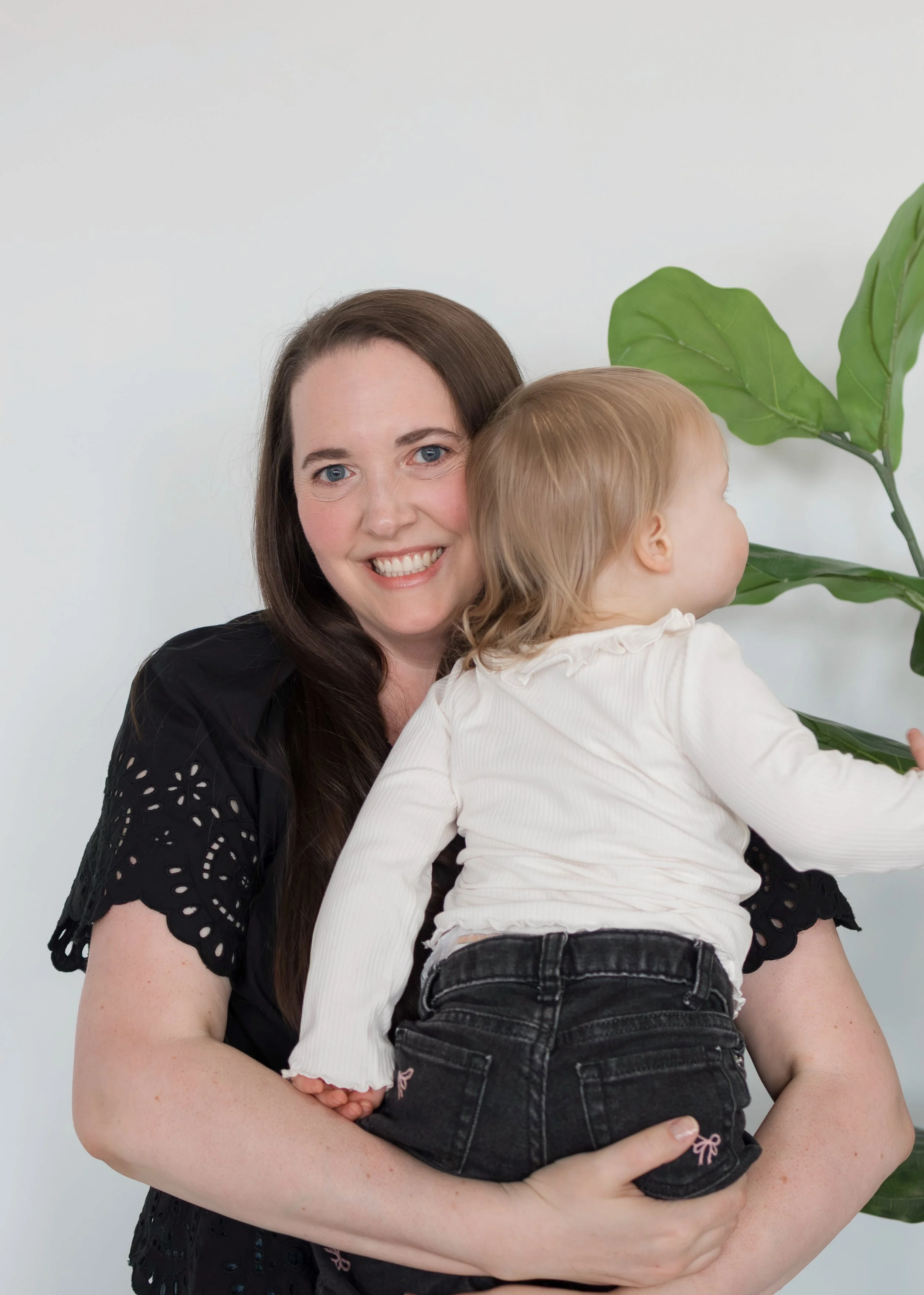 A woman holds a young child, she is facing the camera and smiling. The child is looking over her shoulder