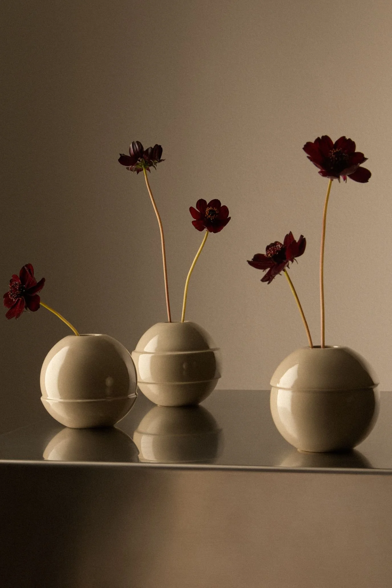 Three rounded Kelly Hoppen x M&S neutral vases holding individual burgundy flower stems.
