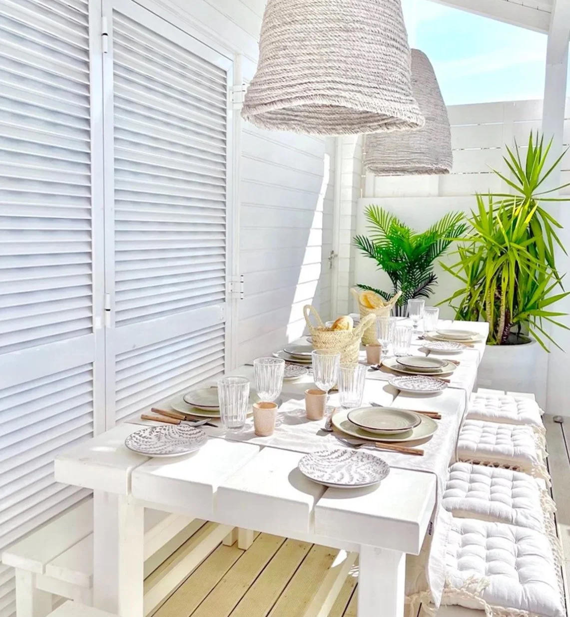 An outdoor white table with tableware, linen runners and glasses with two large rope pendant lights hanging above and potted plants behind