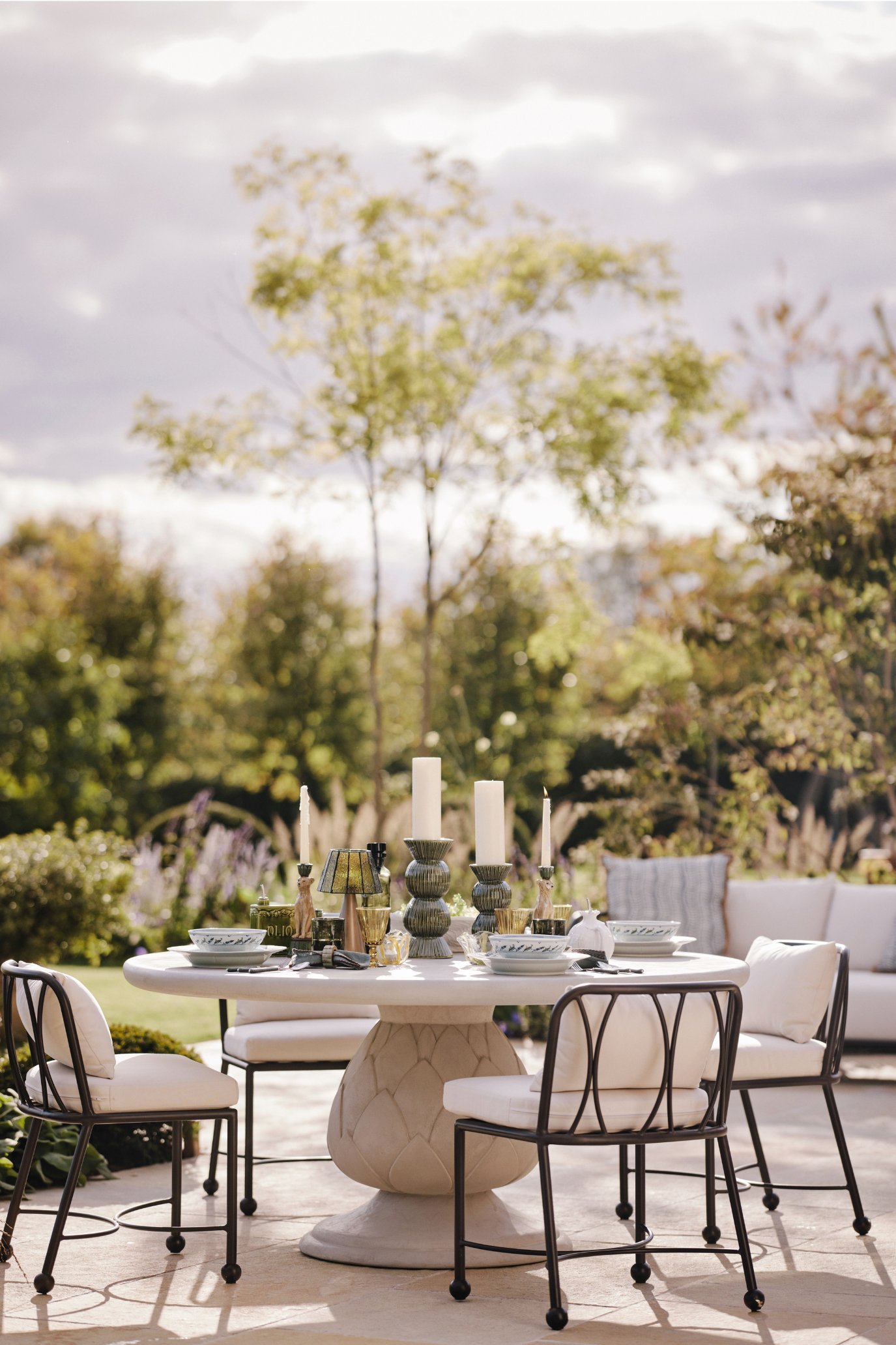 Round outdoor dining table with sculptural artichoke-inspired pedestal base styled with candles and tableware on a sunny terrace.