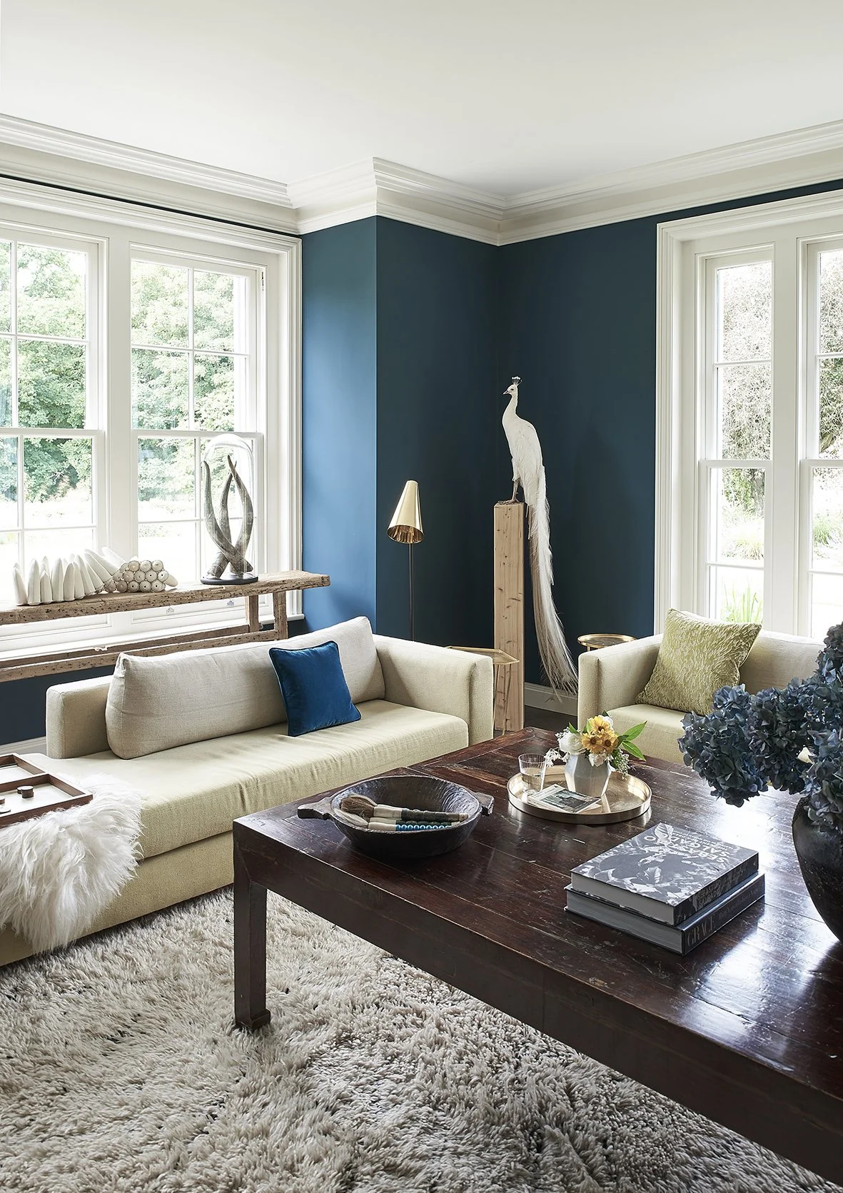 Home decor styling in a blue painted living room with a white peacock on a wooden plinth and  light yellow sofas with blue cushions. a wooden coffee table is decorated with horse brushes, books and flowers.