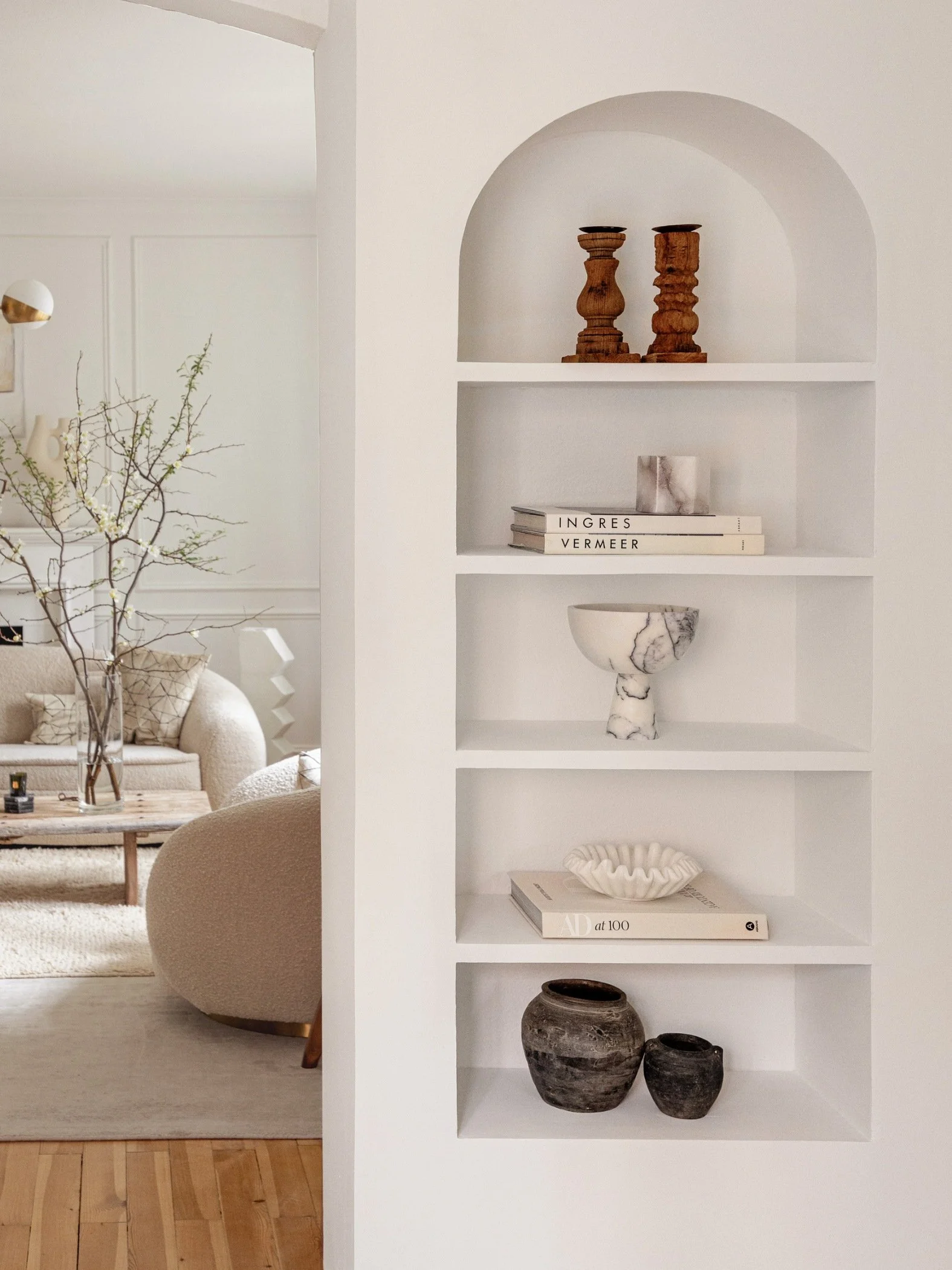 Arched recessed shelving with two amber toned candle sticks on top followed by books, a marble vessel, books with a white dish and two antique pots at the bottom