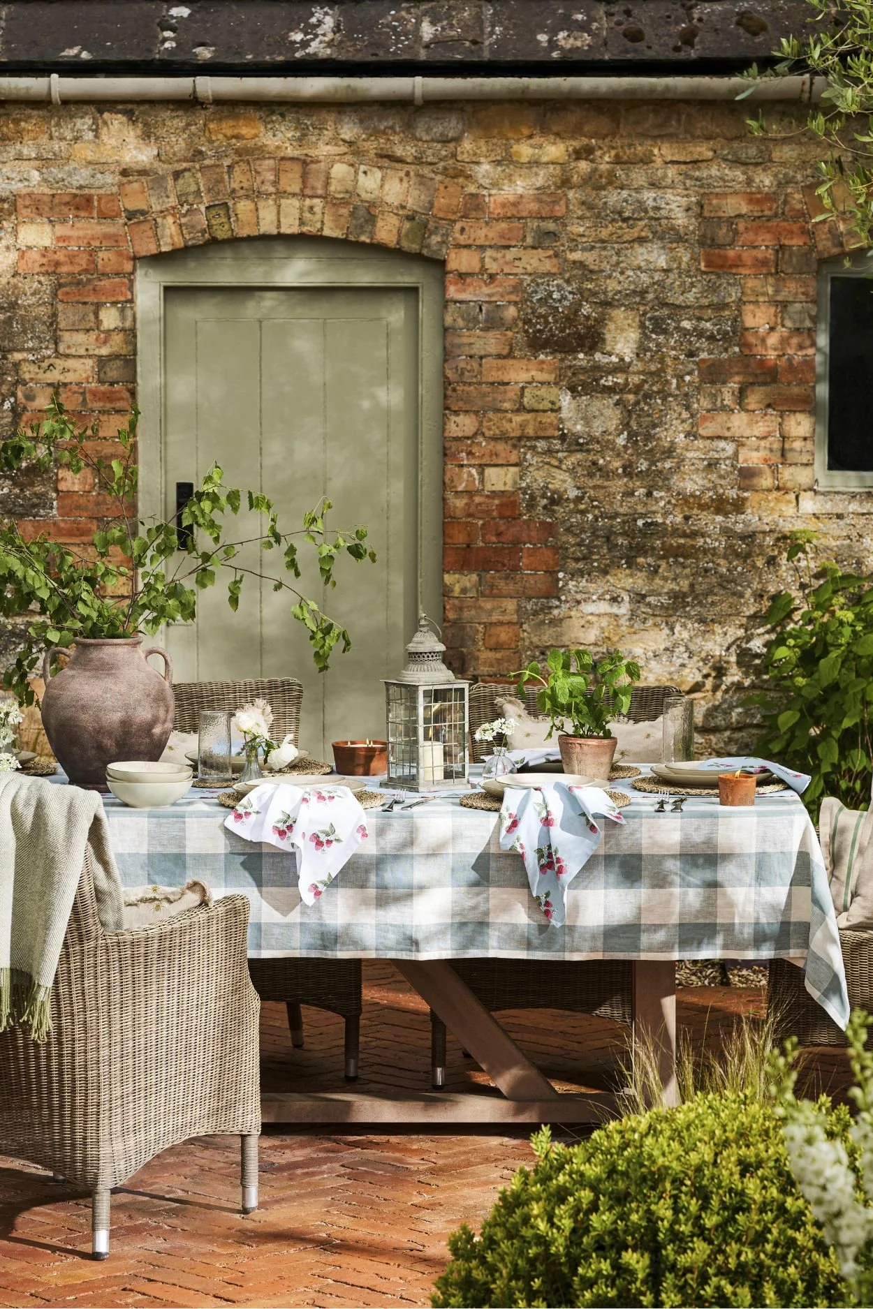 Rustic outdoor dining table styled with a checked tablecloth, floral napkins, lanterns and garden flowers beside a brick wall.