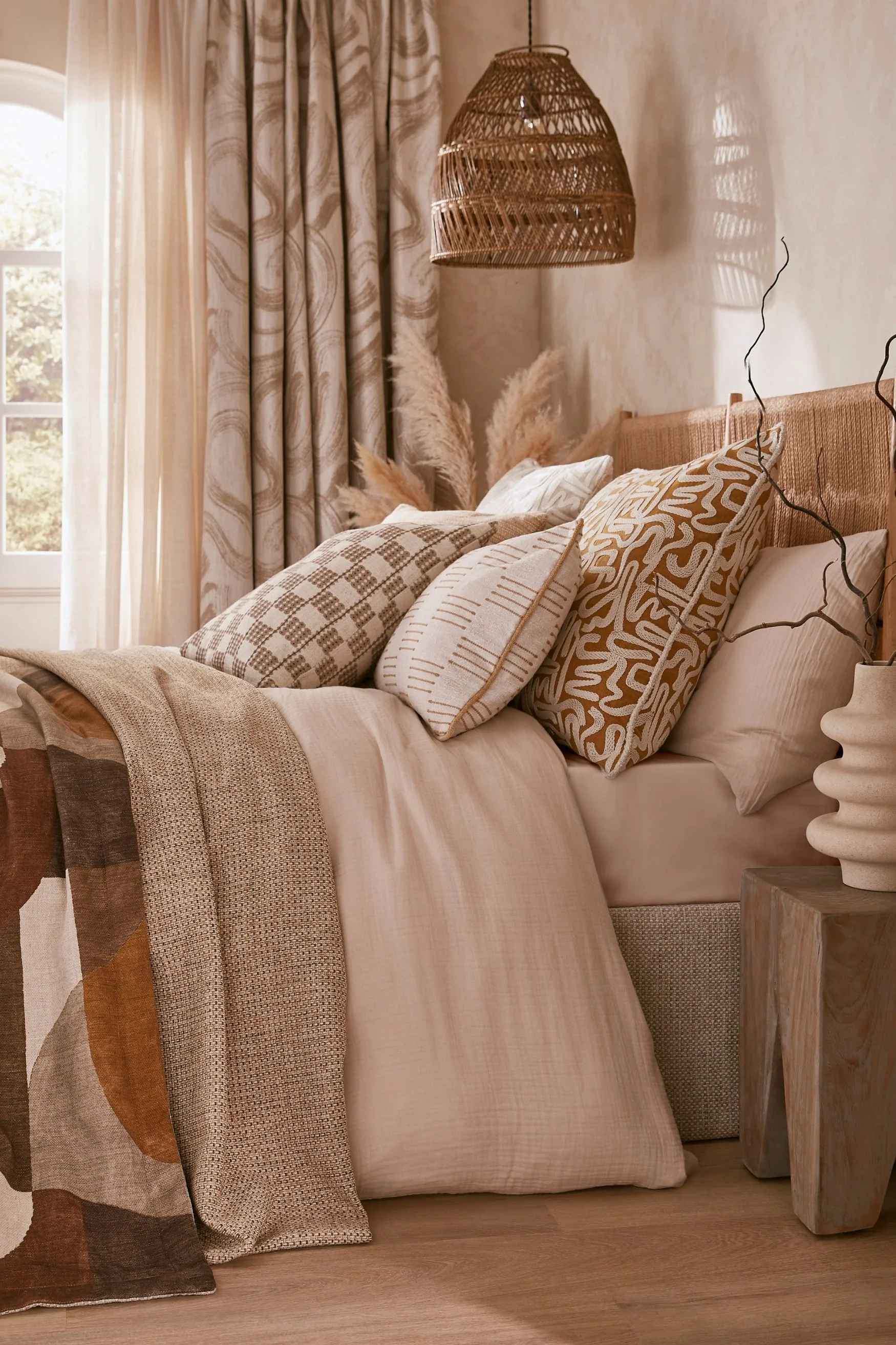 A bedroom with curtains and bedlinen in different soft toned beige printed fabric with a rattan pendant light and rope woven headboard