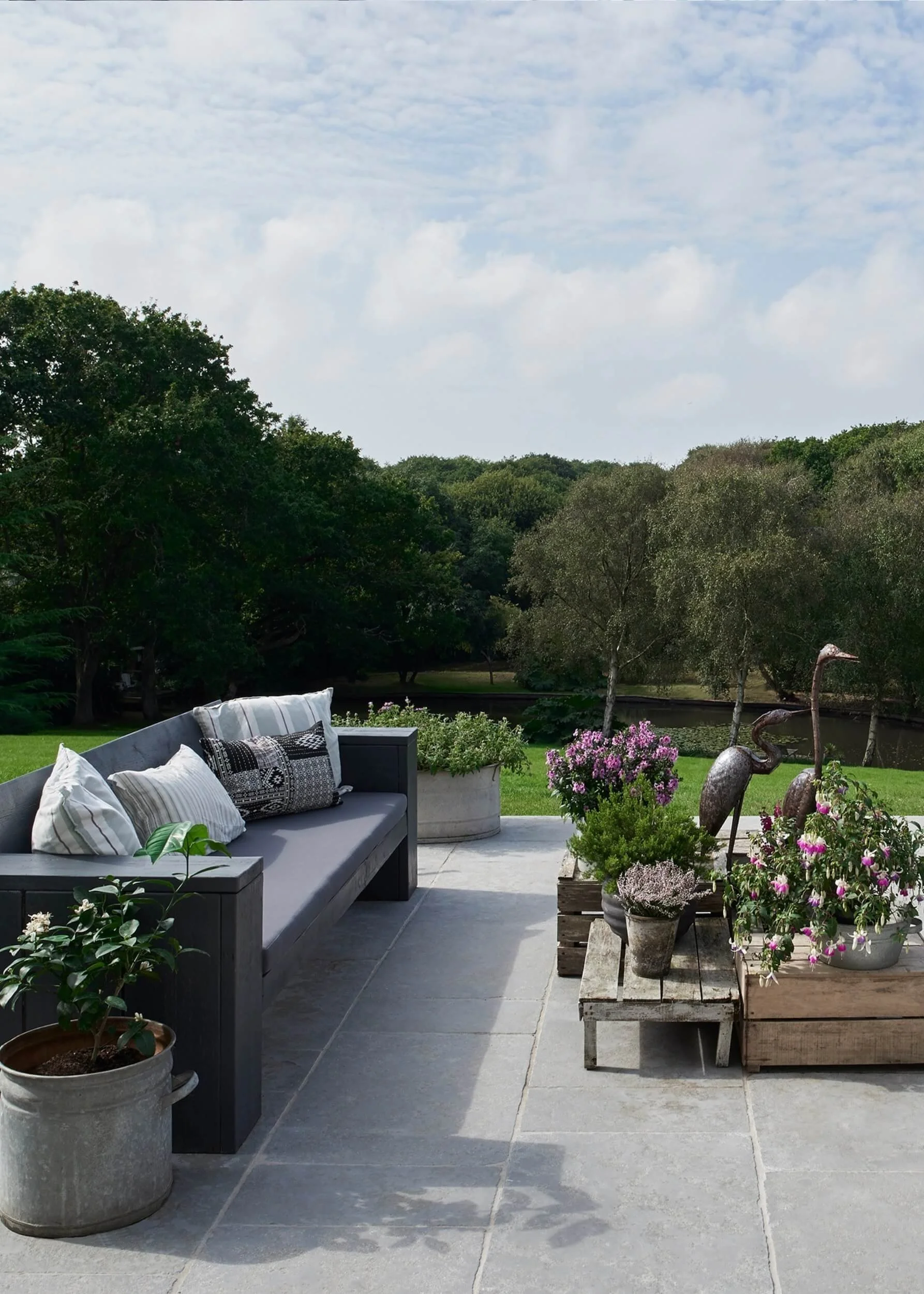 homemade furniture  made from scaffolding planks painted in dark grey with a mixture of grey and white linen cushions, flowers in planters with view of pond.
