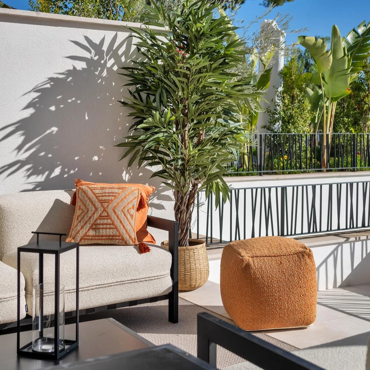 A stylish metal and boucle outdoor sofa styled with burnt orange cushions with a tall plant in a rattan pot next to it