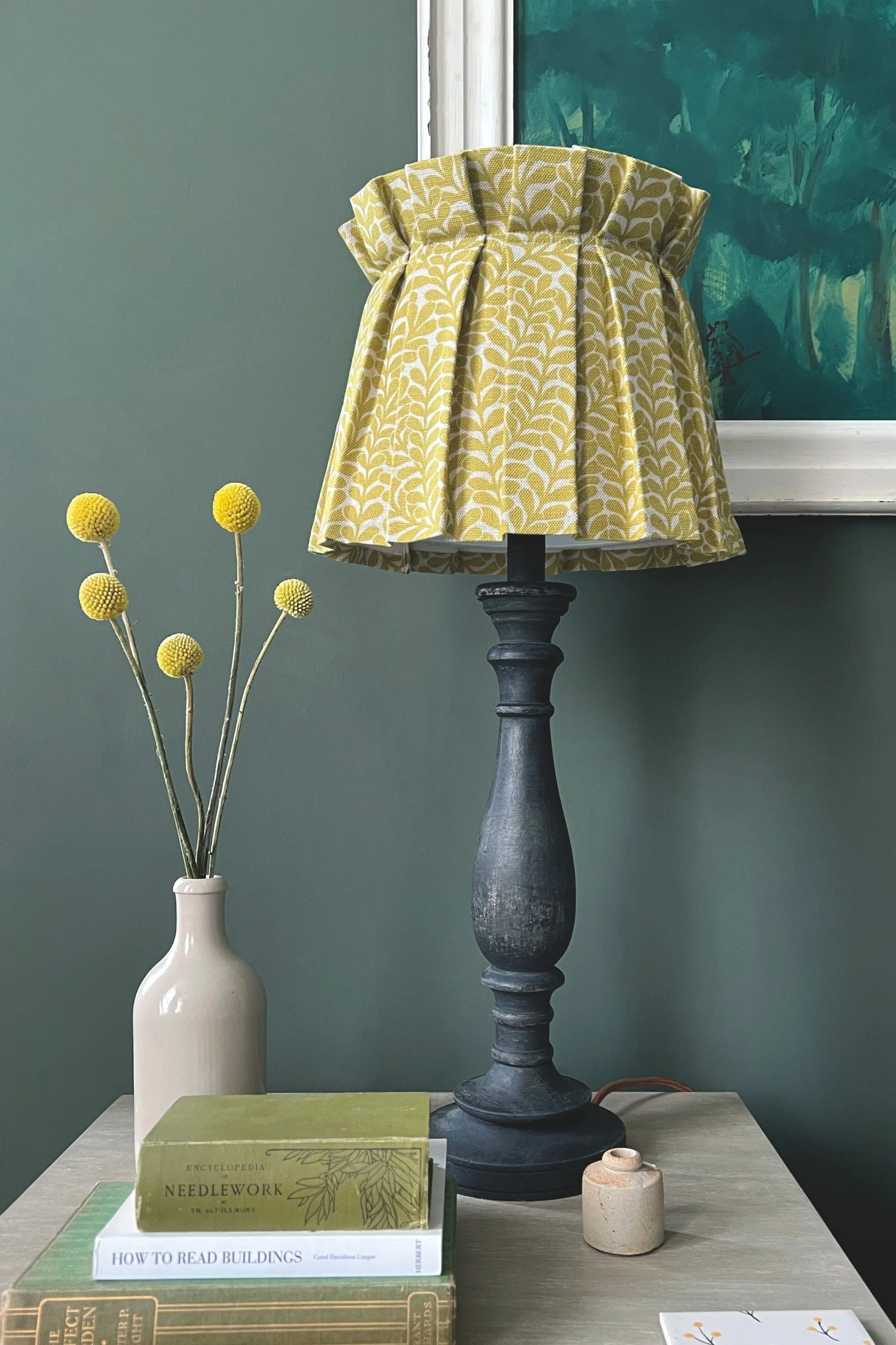 five Billy Button flowers in a white ceramic bottle next to a table lamp with a yellow shade