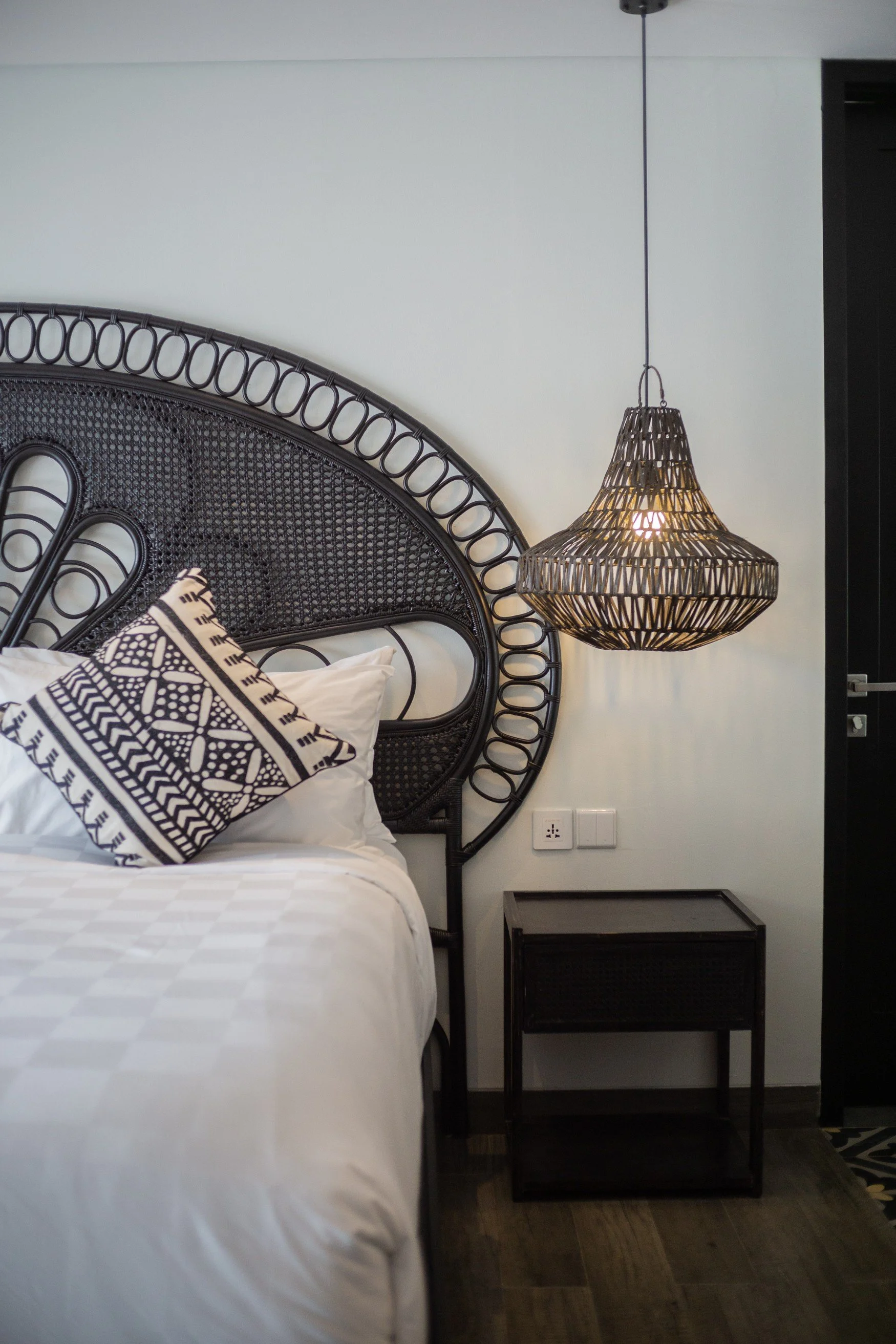 A black fan backed headboard on a bed with white linen and one black and white cushion and a hanging rattan pendant light