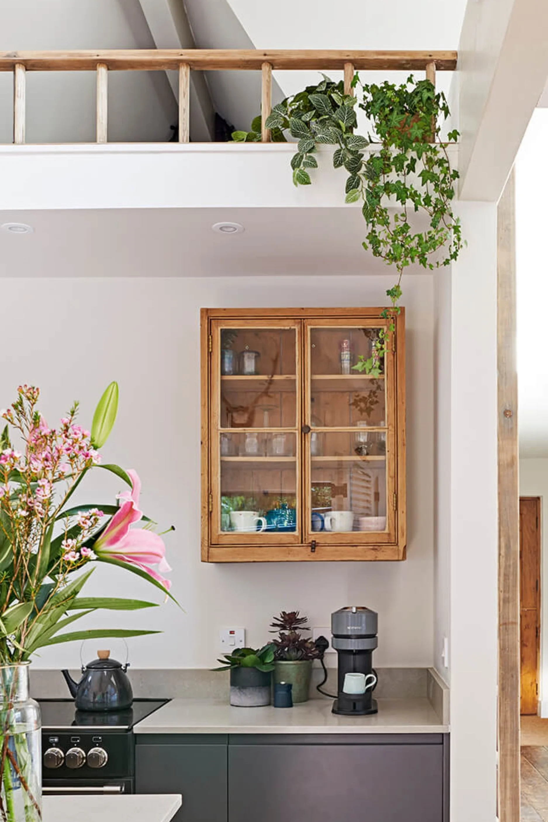 A close up of part of a kitchen with a kettle and coffee maker and a wood, glass fronted cabinet on the wall with glass and crockery in it.