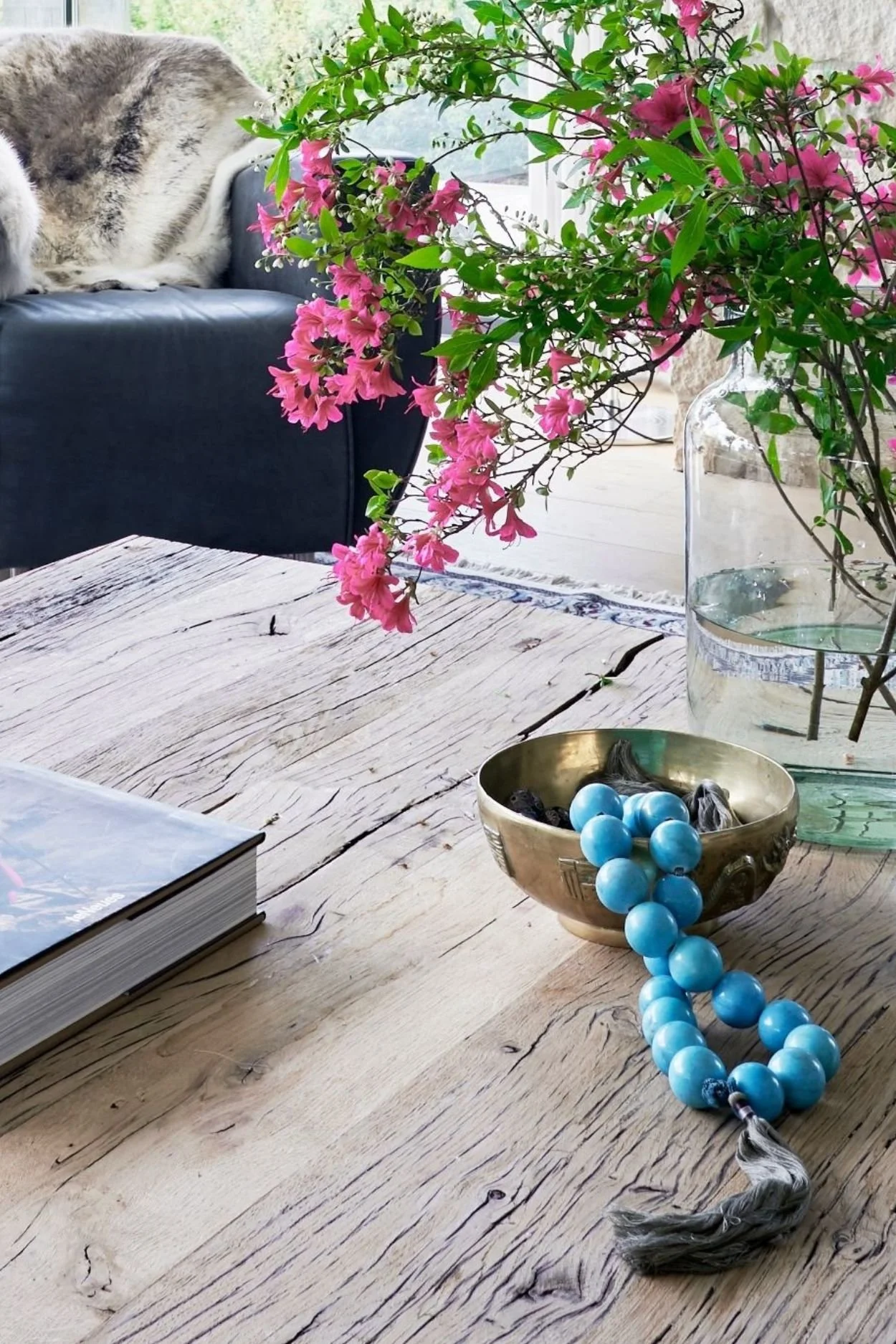 a rustic table with a vase of  pink azaleas and a brass bowl with aqua toned beads