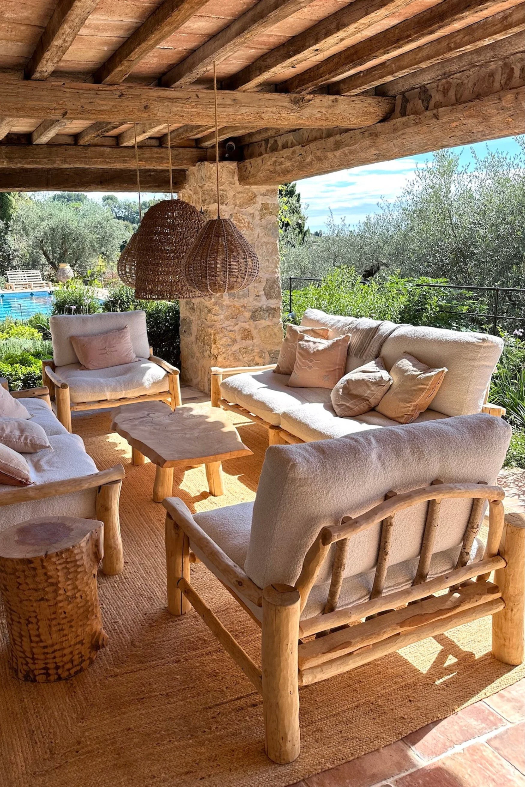Outdoor seating made from driftwood with cream linen cushions and a coffee table. There are three woven pendant lights and a swimming pool in the distance