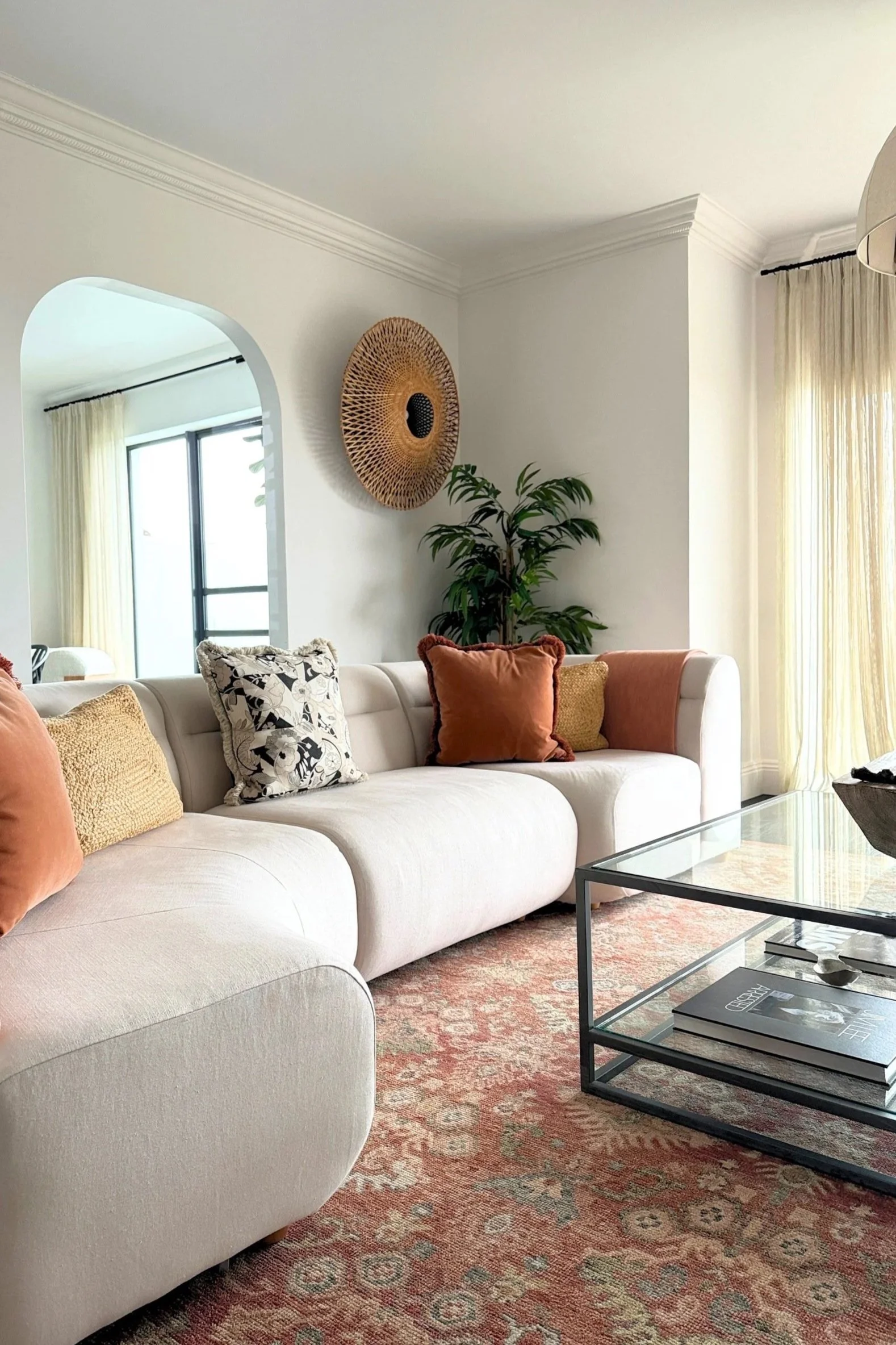An off white sofa with terracotta toned cushions and rug infront of a plant and round rattan wall light