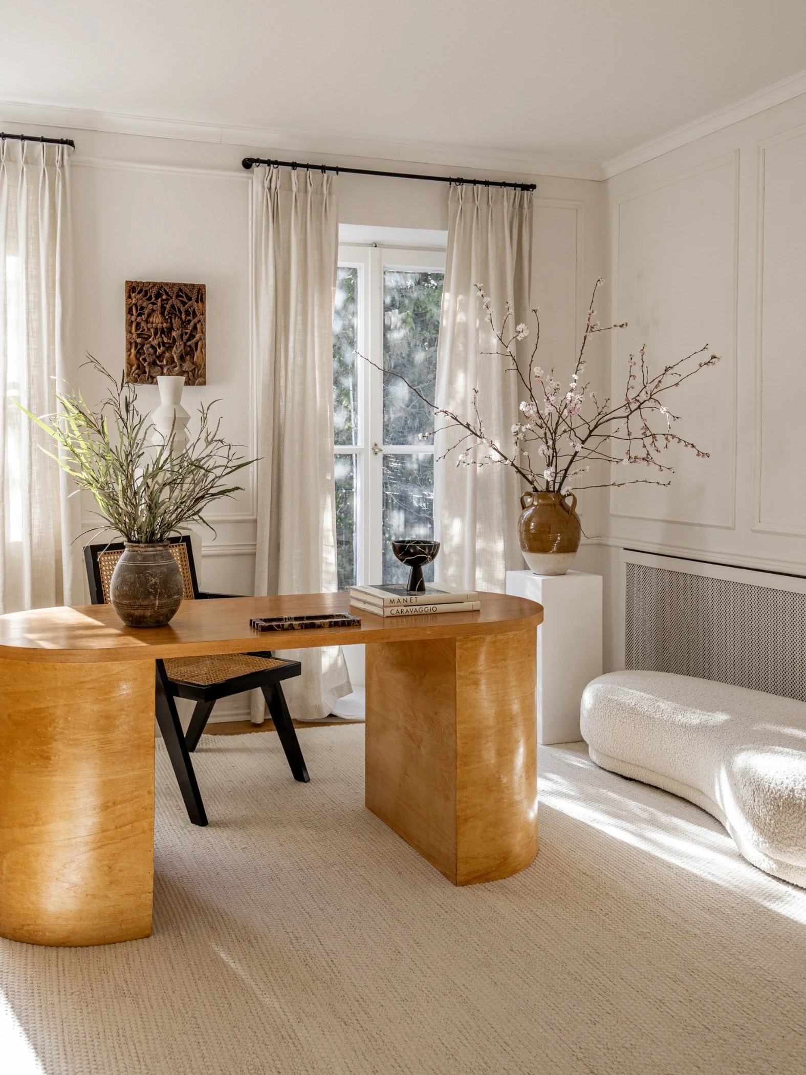 A home office with a curved desk in an amber toned wood, a cream sofa, two vases filled with flowers and grasses and a cane and wood chair