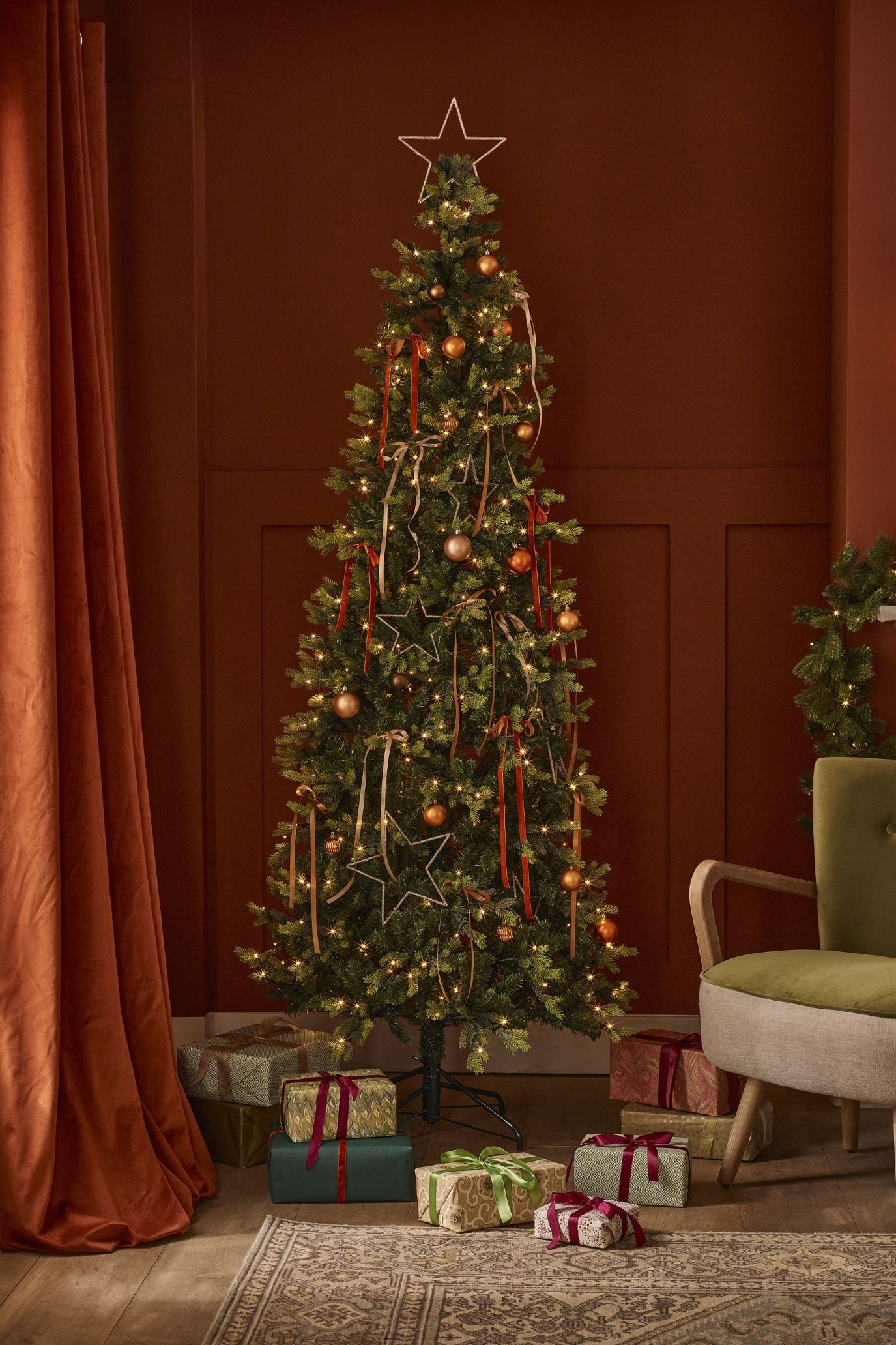 Slender Christmas tree with metallic baubles, delicate ribbon bows and open star ornaments against a deep red wall.