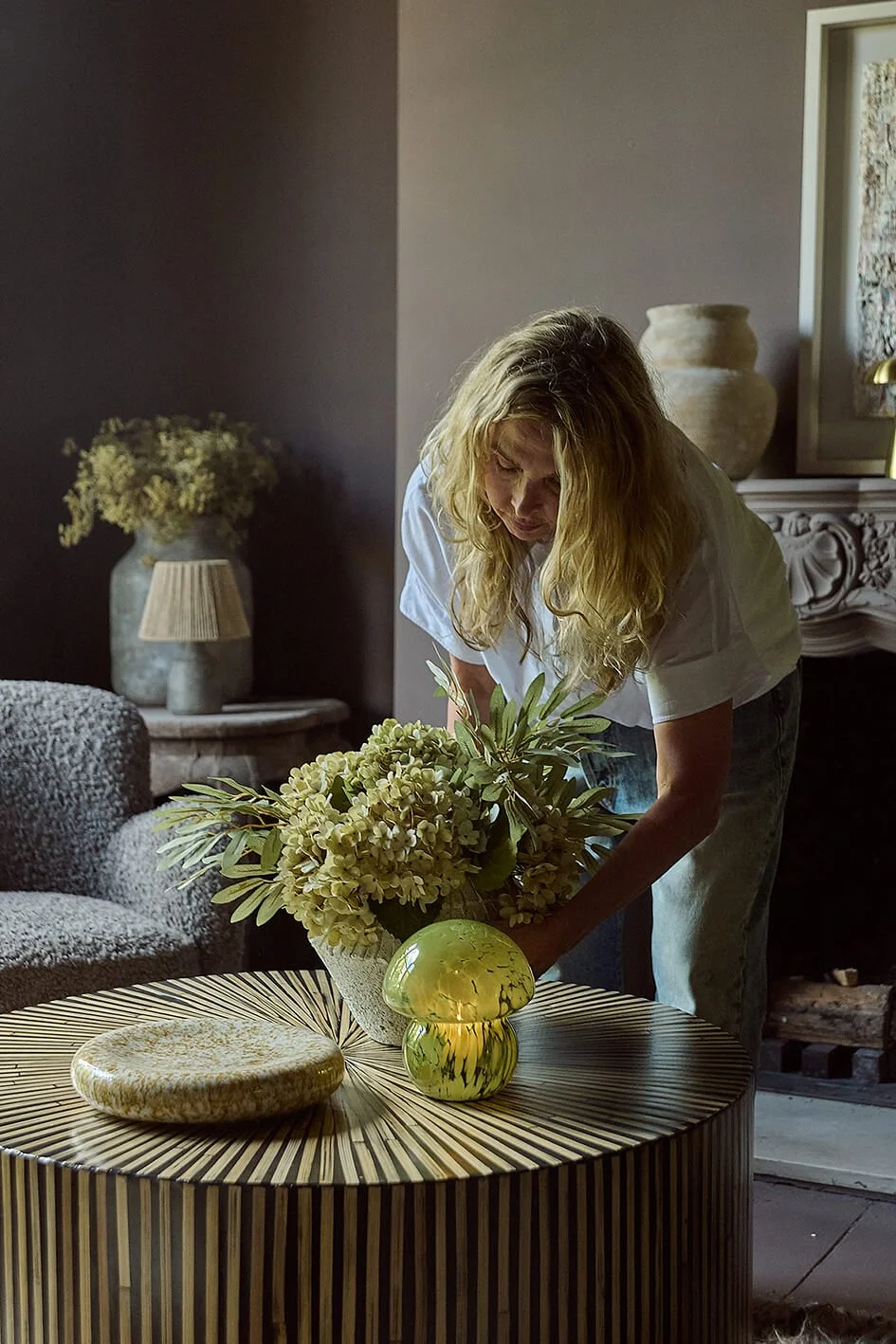 interior Designer Abigail Ahern sitting on a fur chair next to  a black round sculptured table with a cream lamp on it with recessed shelves behind