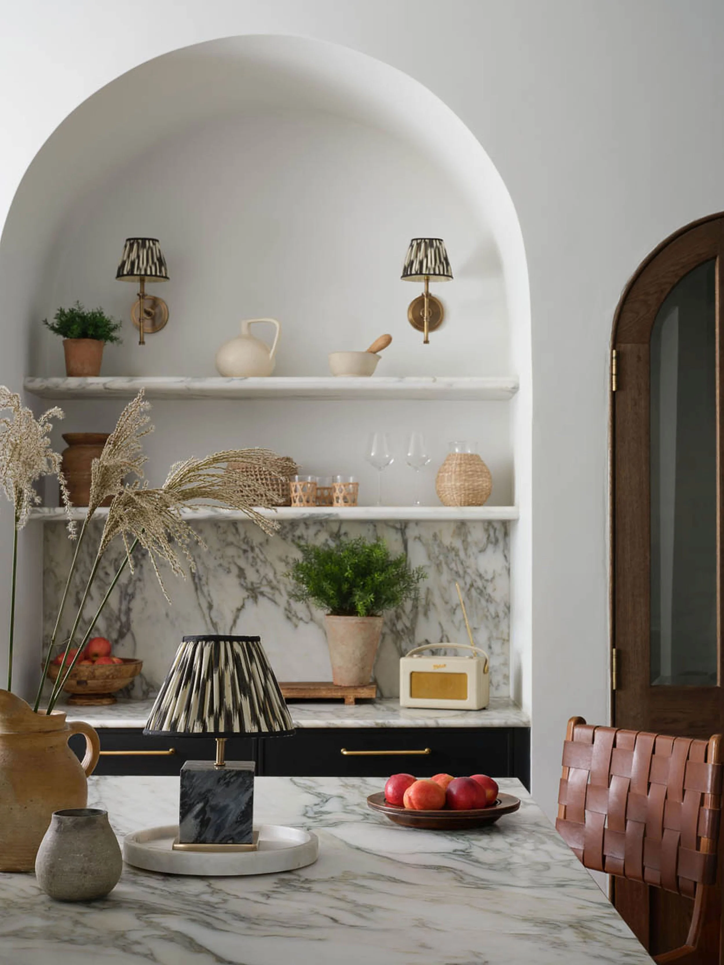 An arch with two shelves and wall lights with a kitchen island in front styled with a lamp, stone jugs and a bowl of red apples