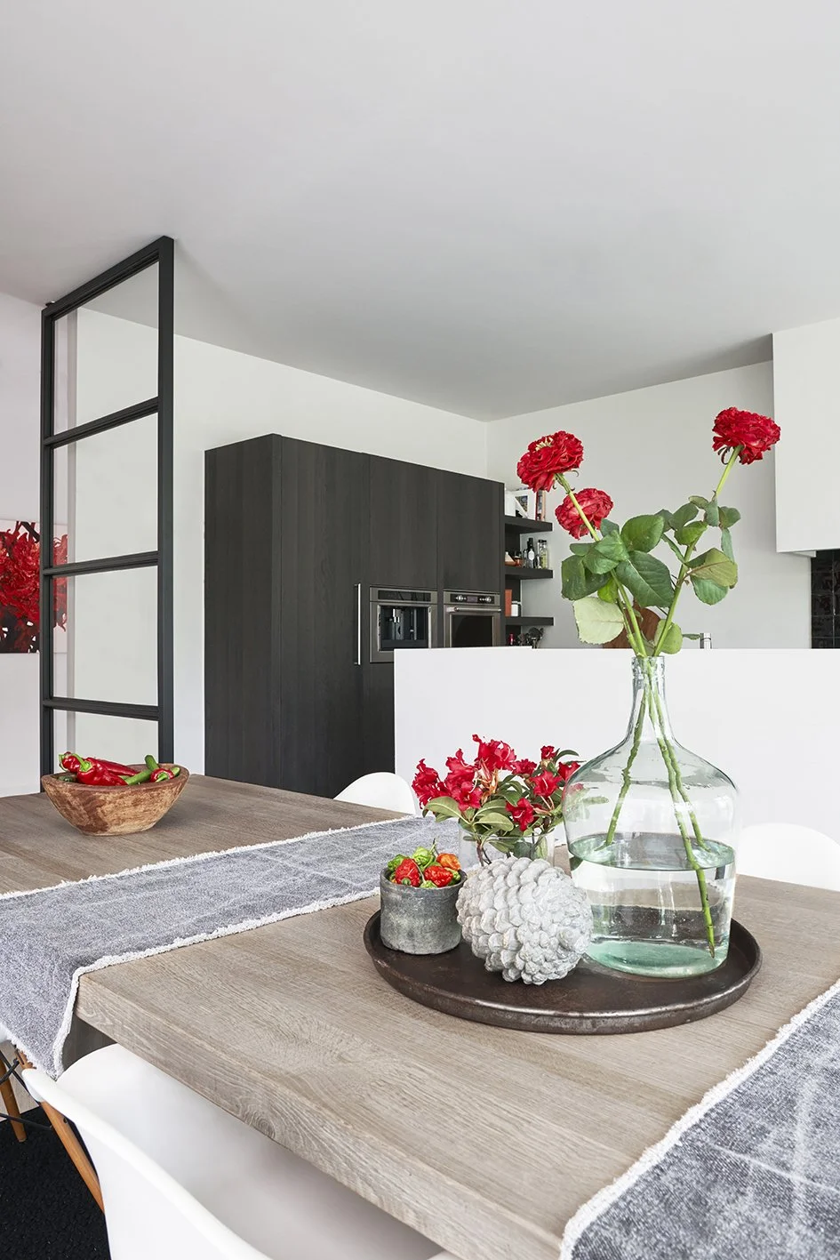 a stylish metal tray with a vintage vase with red flowers a pot with chilli peppers and decorative acorn on a wooden table with two grey  table runners and a wooden bowl with red peppers.