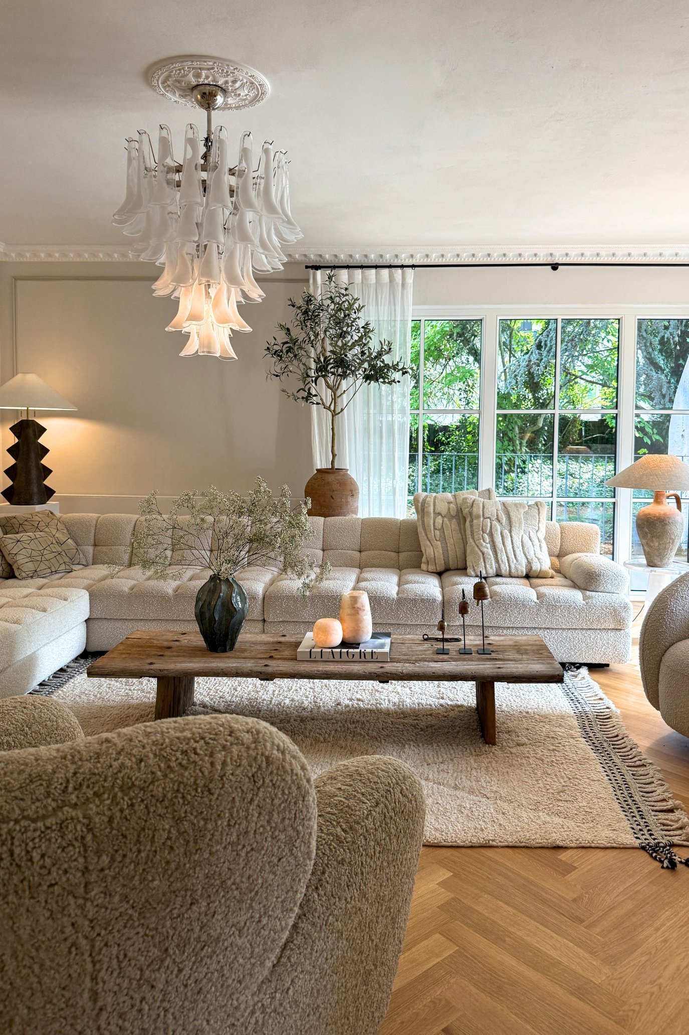 A living room with a large cream boucle corner sofa , a chandelier, a cream rug with a rustic wooden tables with home decor on it.