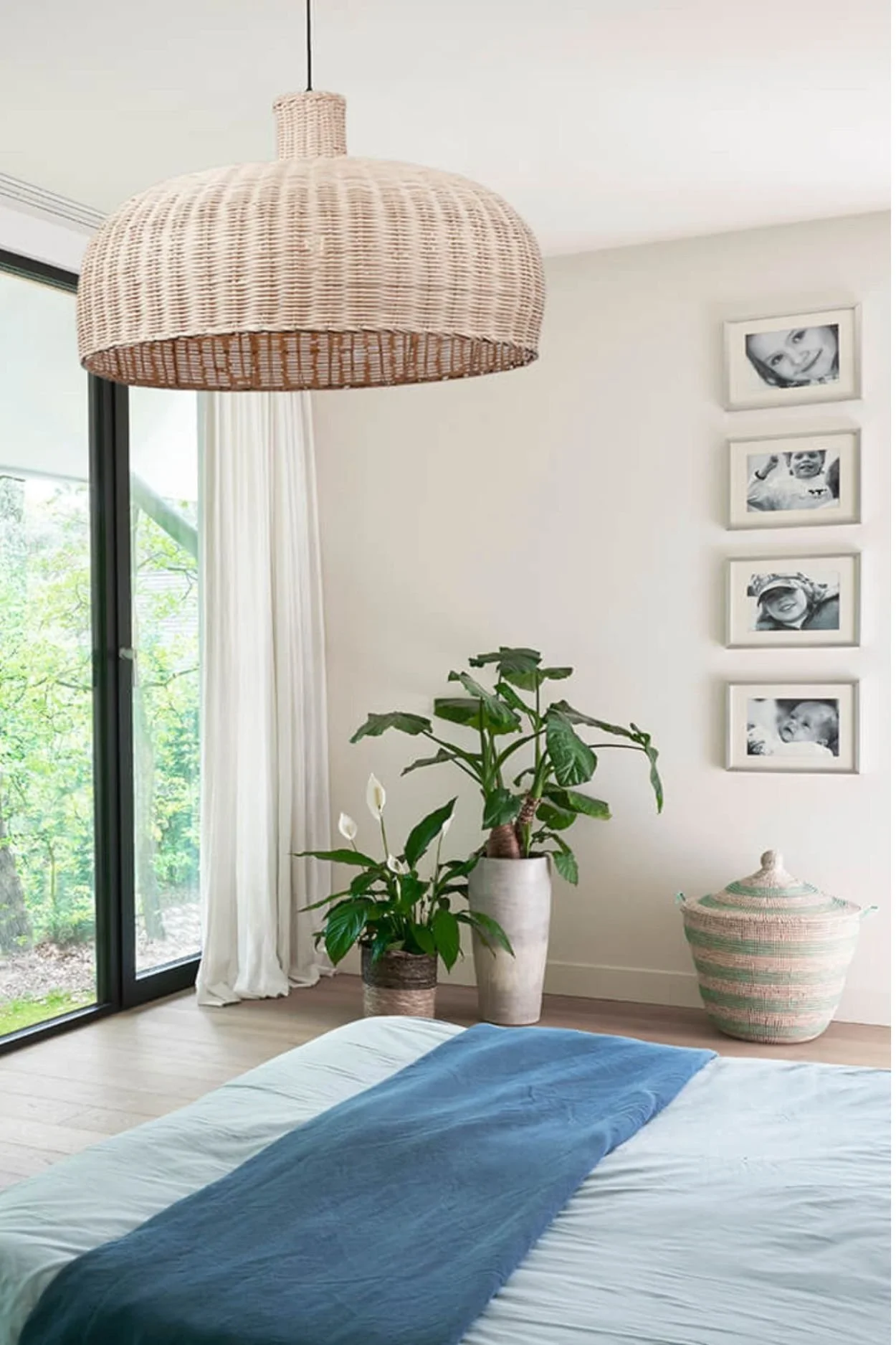 A bedroom with aqua-toned bed line a rattan pendant light, laundry basket and two plants in pots on the floor