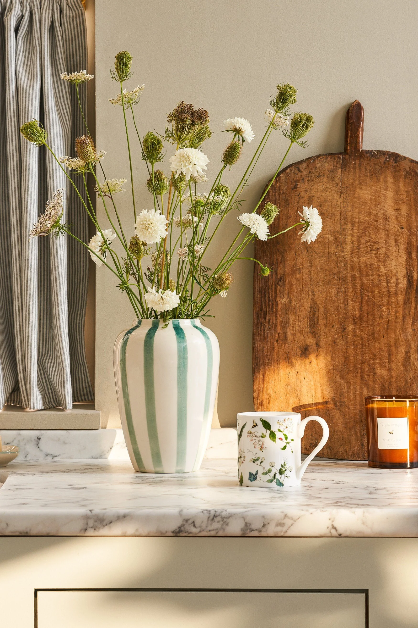 A large vintage wooden chopping board on a kitchen work top next to a white. and green striped vase with white flowers