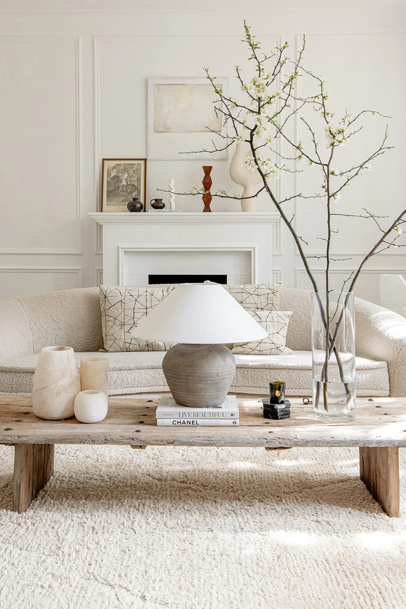 A cream curved sofa in front of a white fireplace and a rustic wooden coffee table with a lamp and white vase with blossom in it