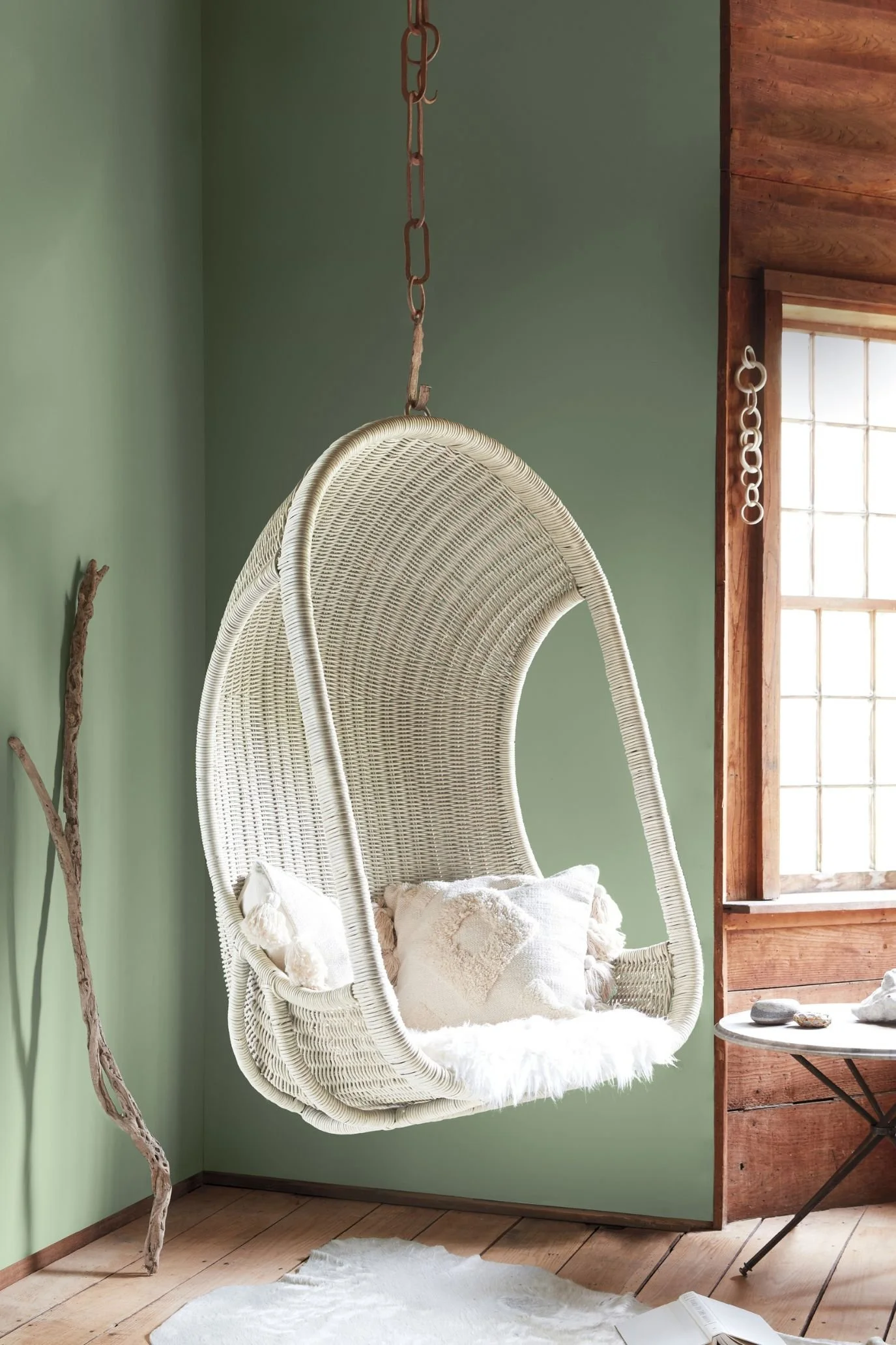 White woven hanging chair with textured cream cushions against soft green walls beside a timber-framed window.