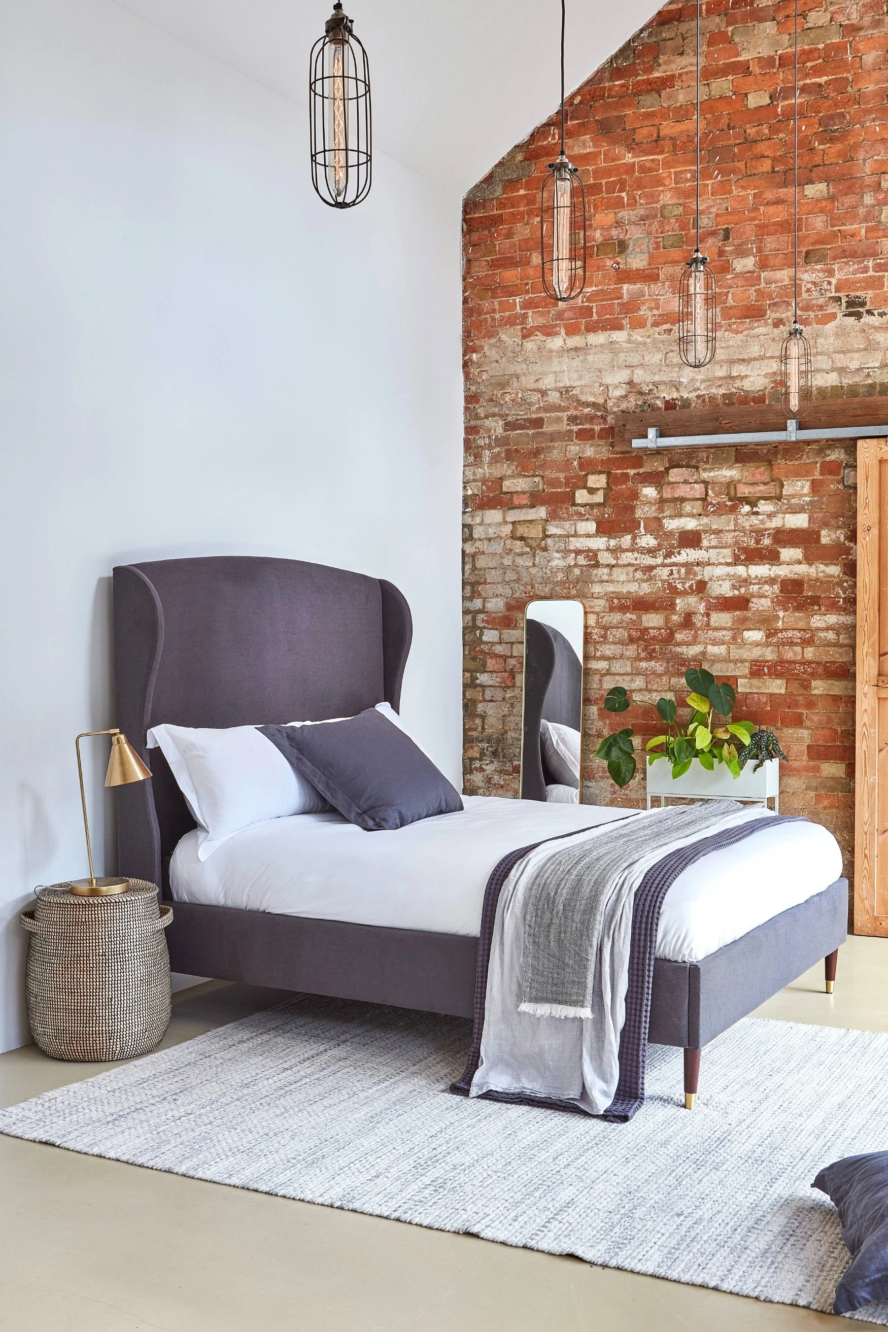 A bed close to an exposed brick wall with a mauve velvet headboard, cushions and throws with a rattan basket and lamp next to the bed and white rug underneath