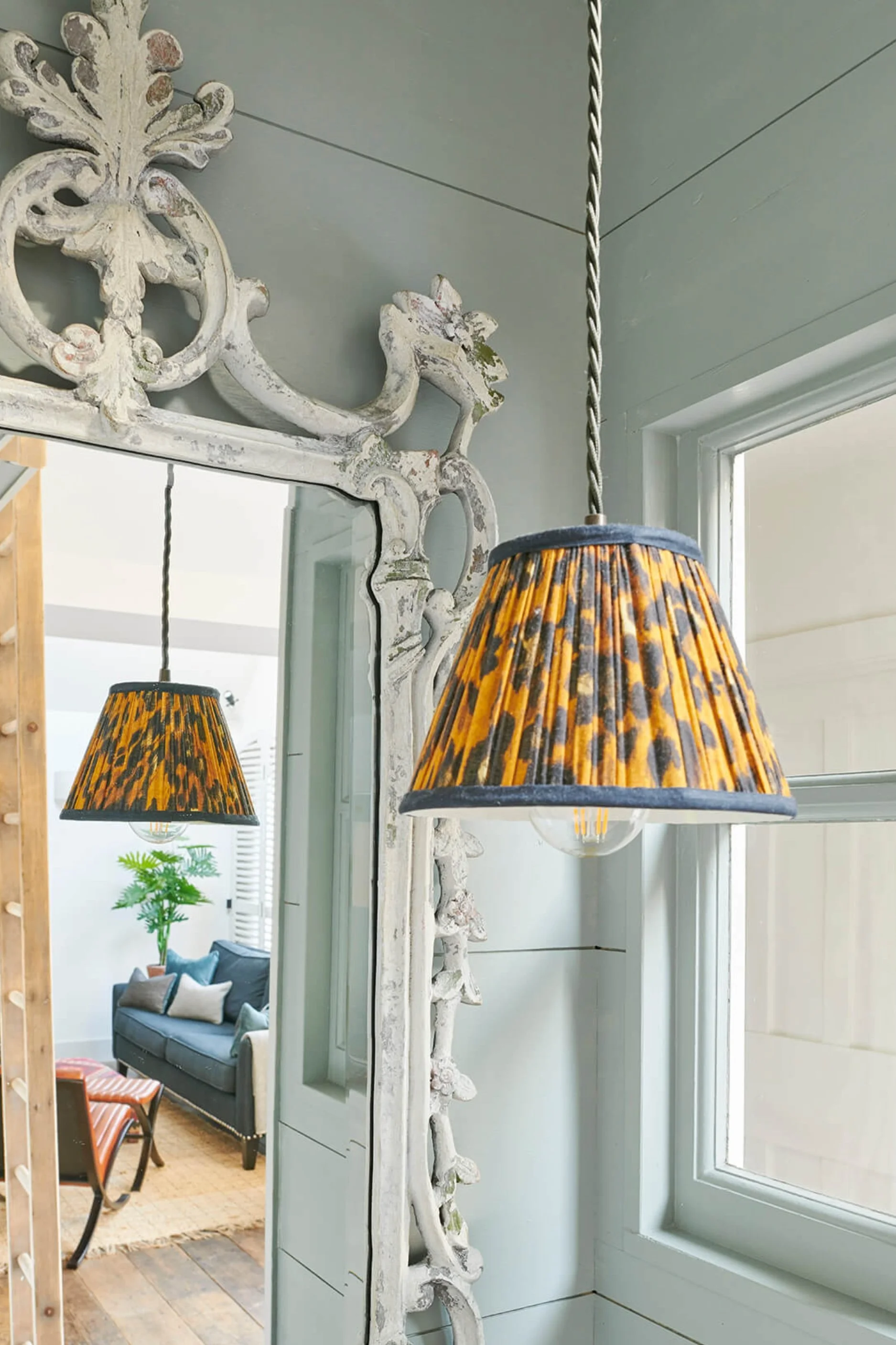 a white ornate mirror with a view to a sofa. A hanging pendant in an animal print is infront of the mirror. You can also see part odf a ladder in the mirror