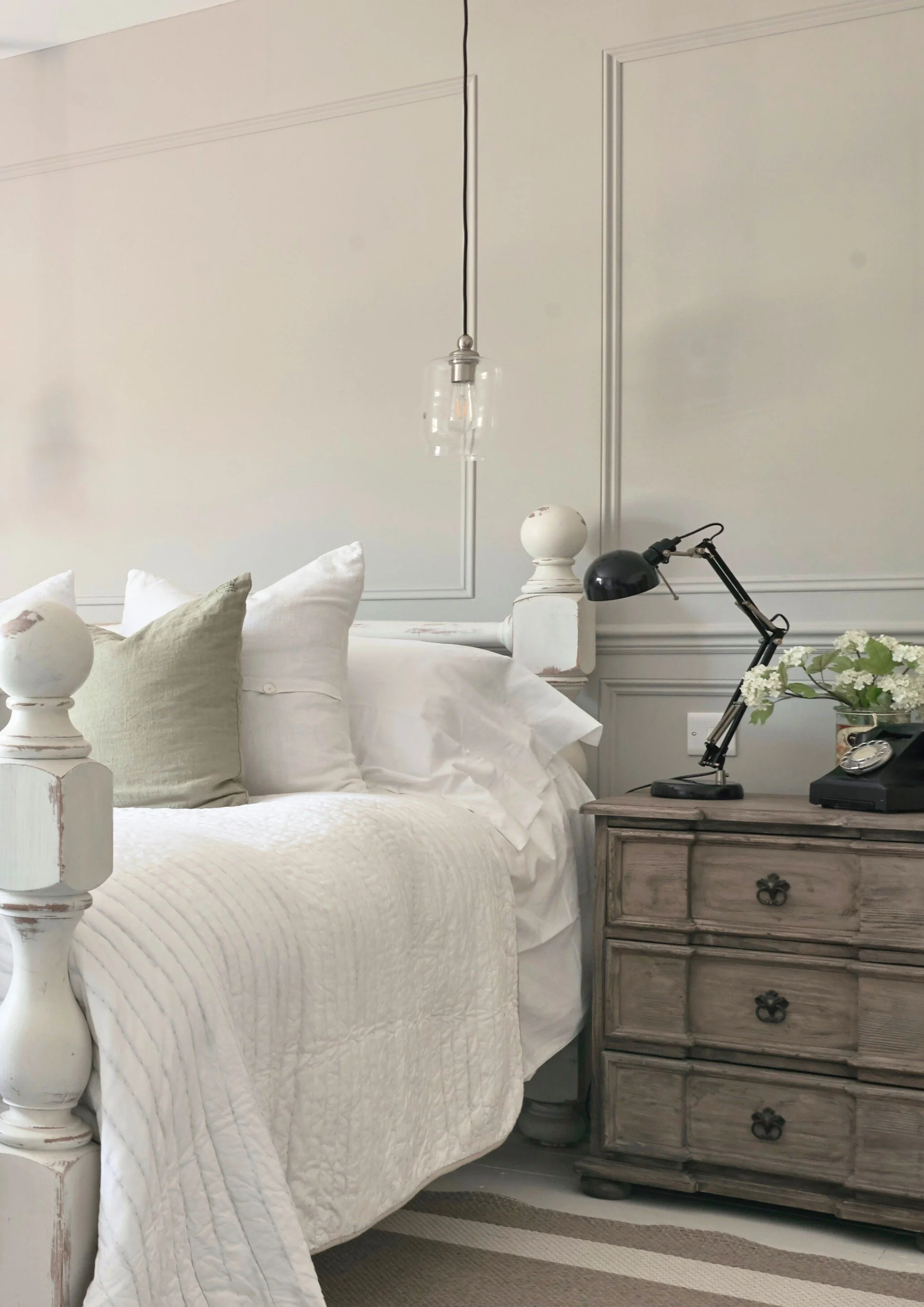 A close up of a bed in white linen with a green cushion next to a vintage chest with a black anglepoise light and retro telephone