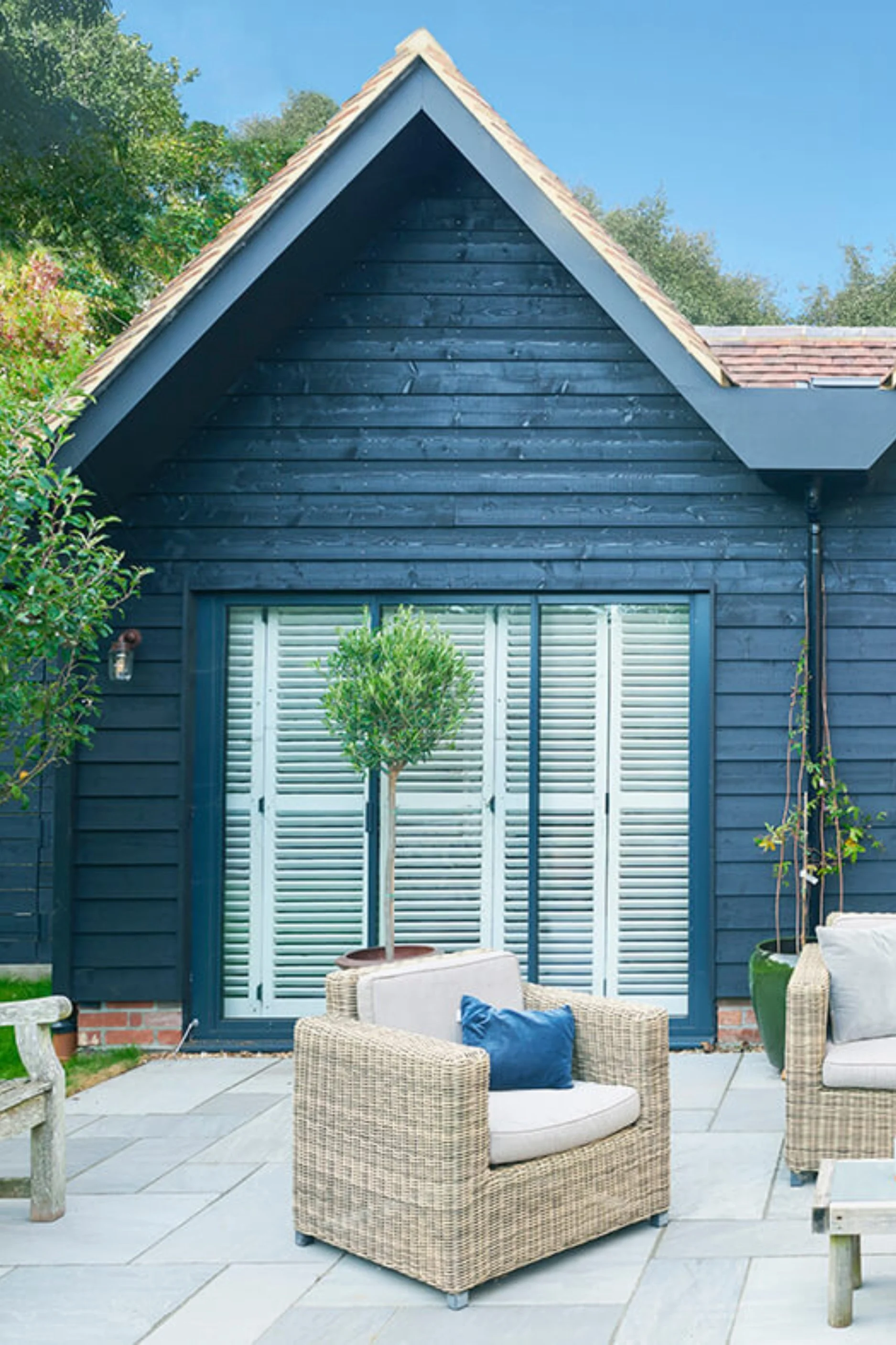 The exterior of a garage conversion with a seating area infront of a building clad in dark wood with a terracotta tiles pointed roof