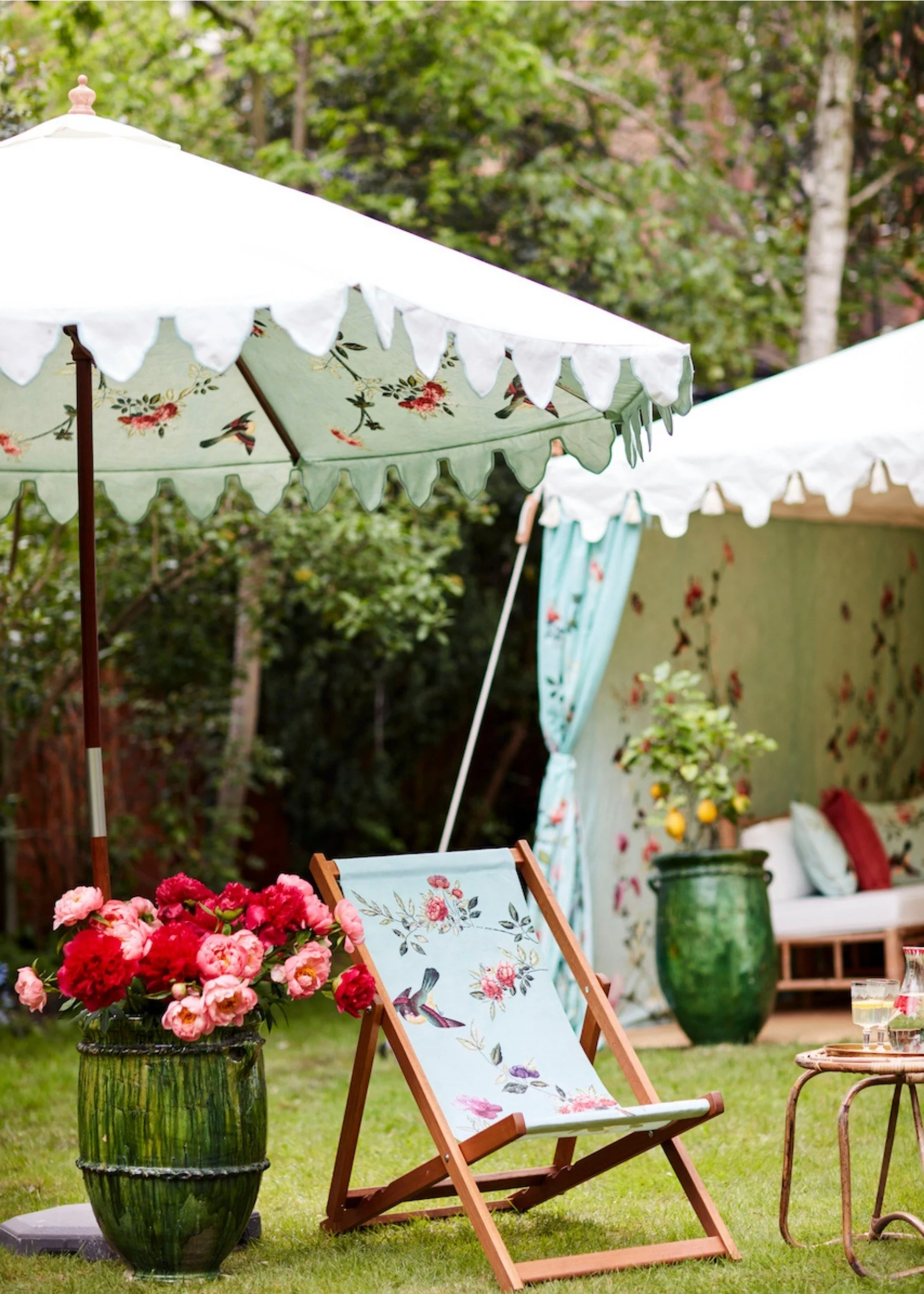 a parasol, and deckchair with a floral design next to a tub filled with pink and red flowers with a marque in the background