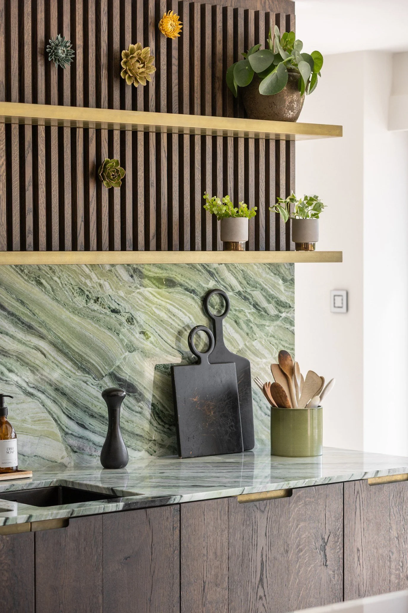 A designer-style kitchen with green marble cplashback and countertop and dark wood cabinetry. Behind two metallic shelves styled with plants is a wooden fluted wall