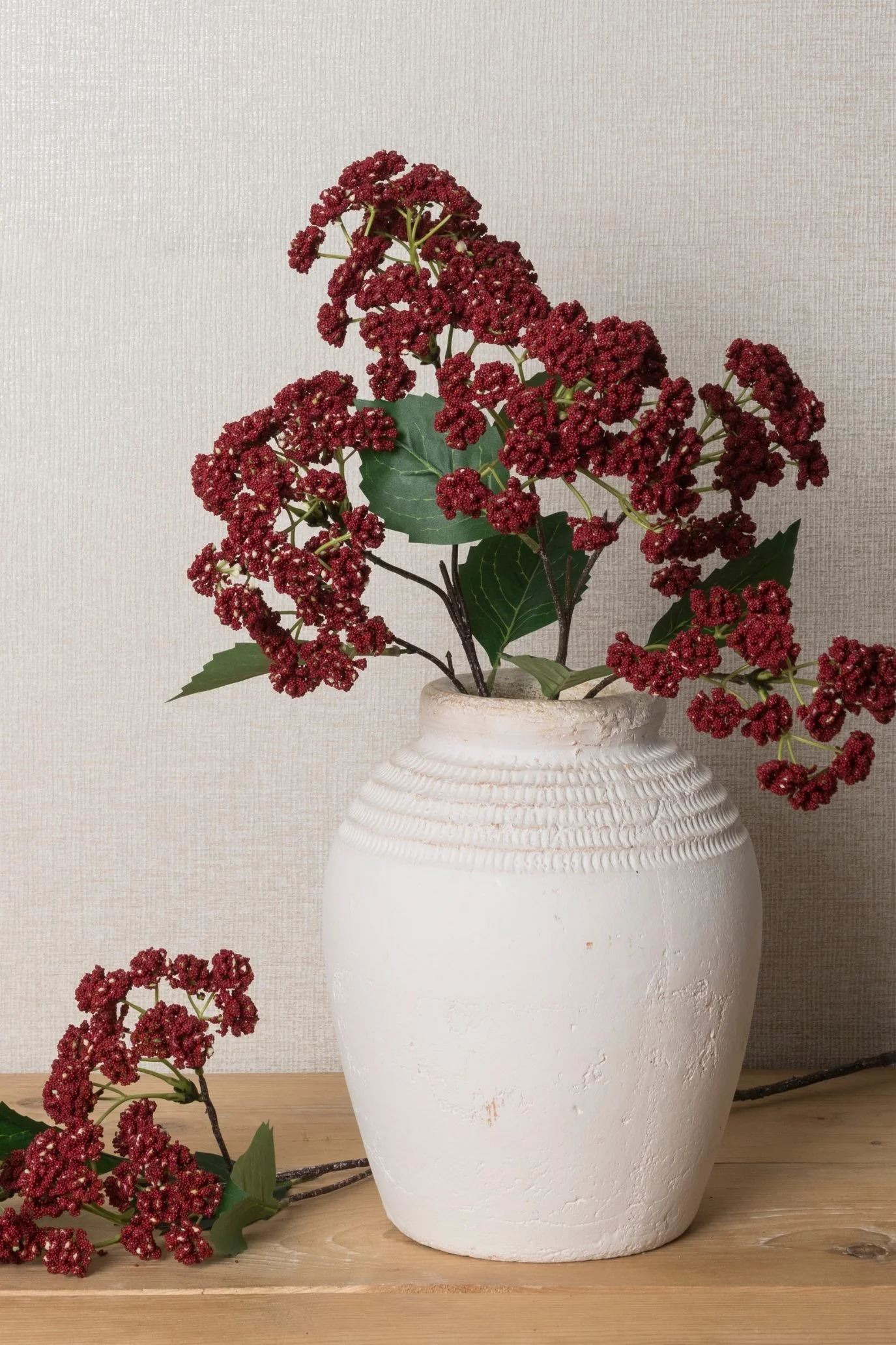 Burgundy faux flowers arranged in a textured white ceramic vase.