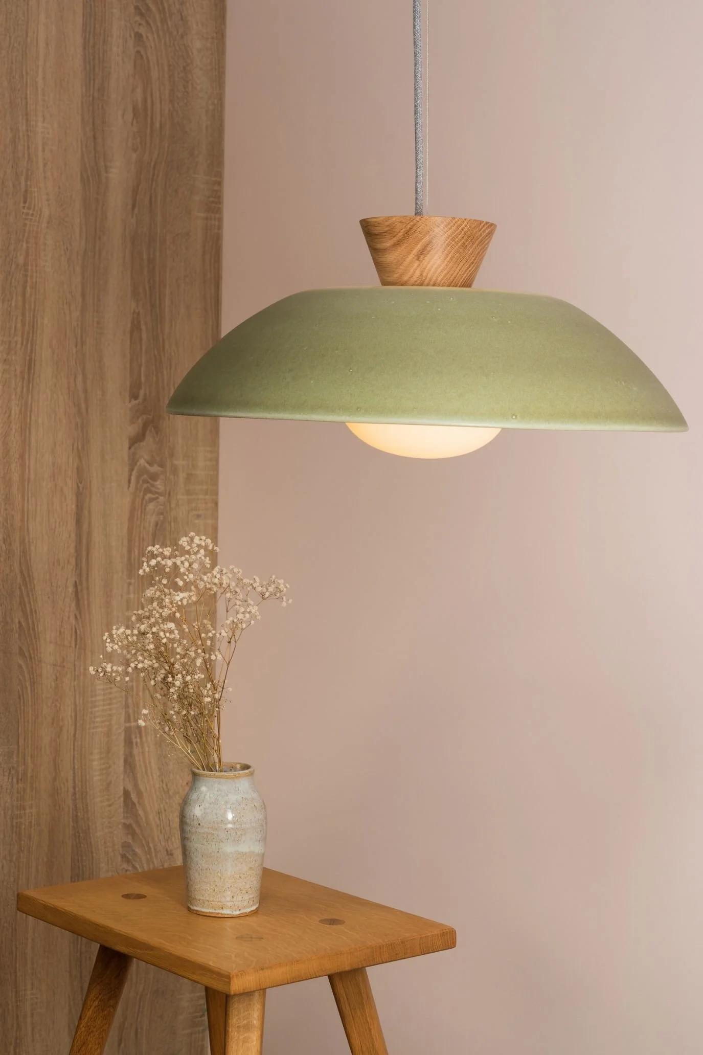 Wide green dome pendant light with a wooden detail above a small wooden table against a pale pink wall.
