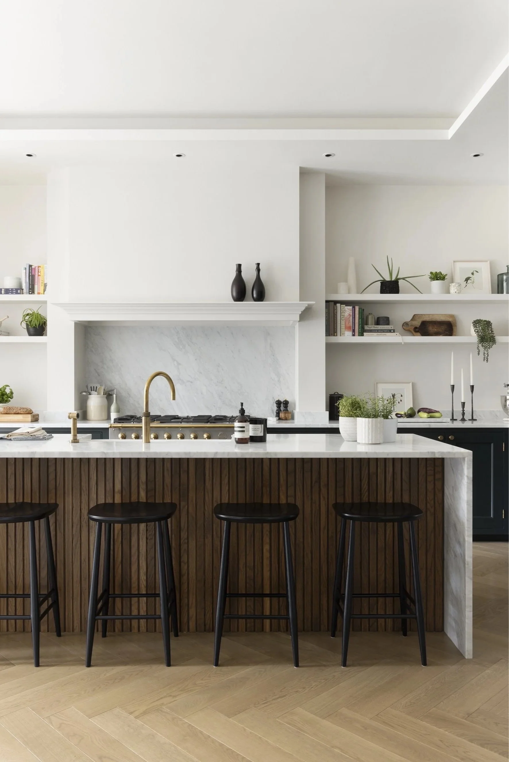 A kitchen with four black stools by a kitchen island and white shelves either side of the hob.