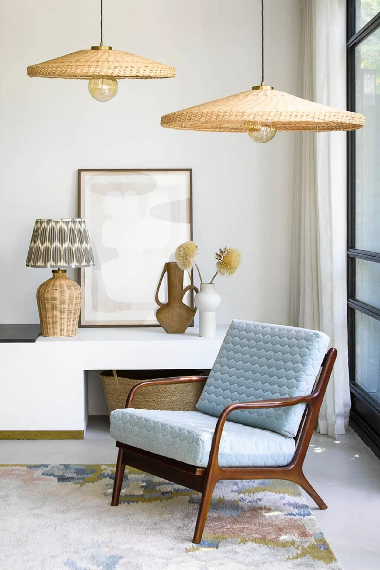 two wide rattan pendant lights above a dark wood chair with light blue upholstery with a rattan lamp, framed art and two vases on a built in surface