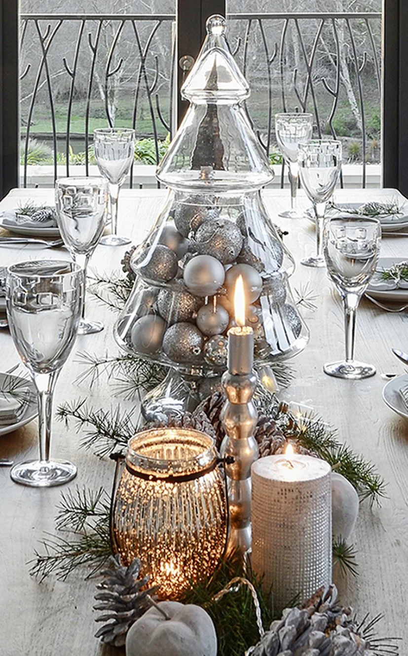 A table laid for Christmas dinner with a glass of christmas tree full of silver baubles and candles infront of it