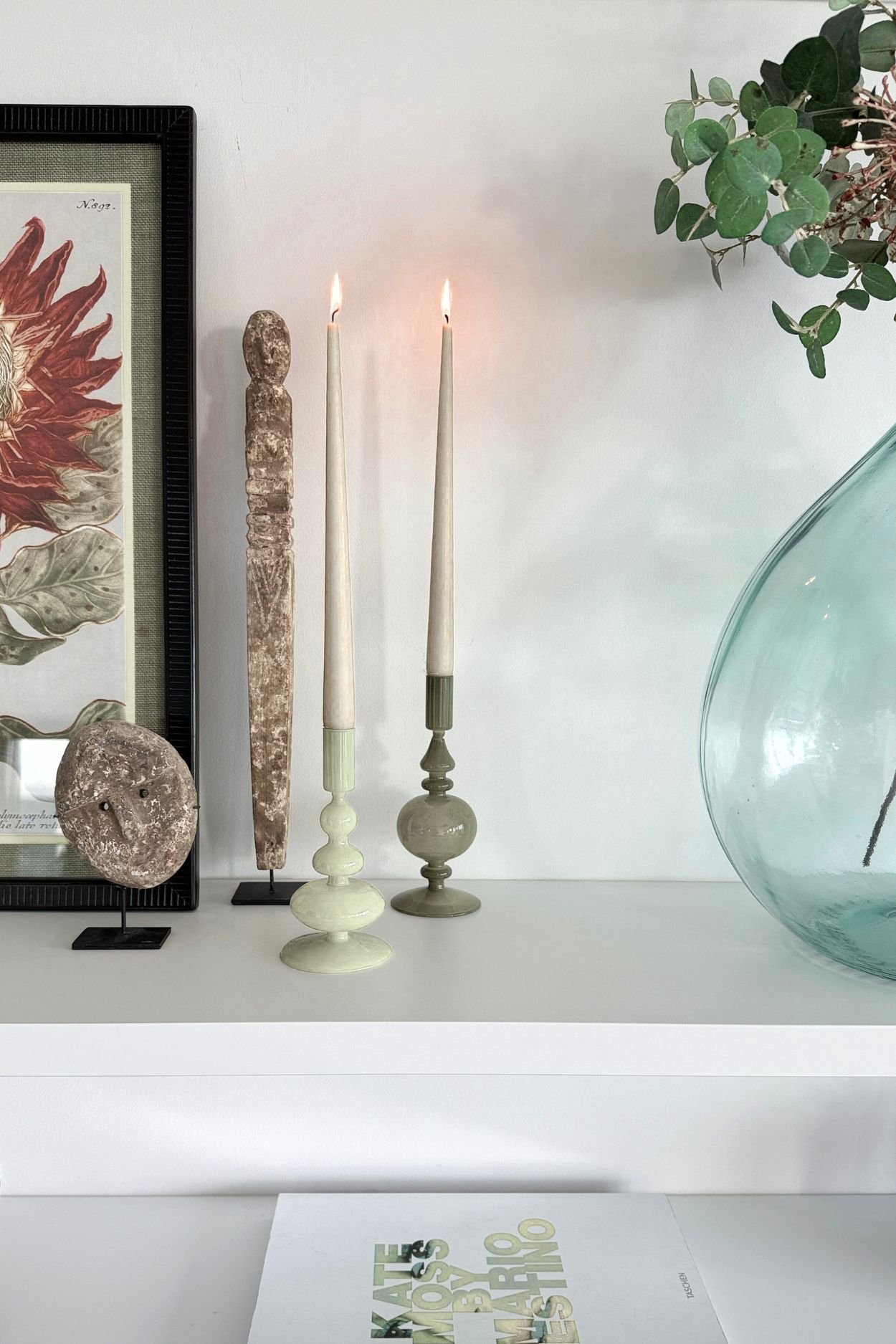 Two shelves one with green candle holders and two wooden characters next to a framed sunflower with a book on the bottom shelf and glass vase with greenery next to the candles