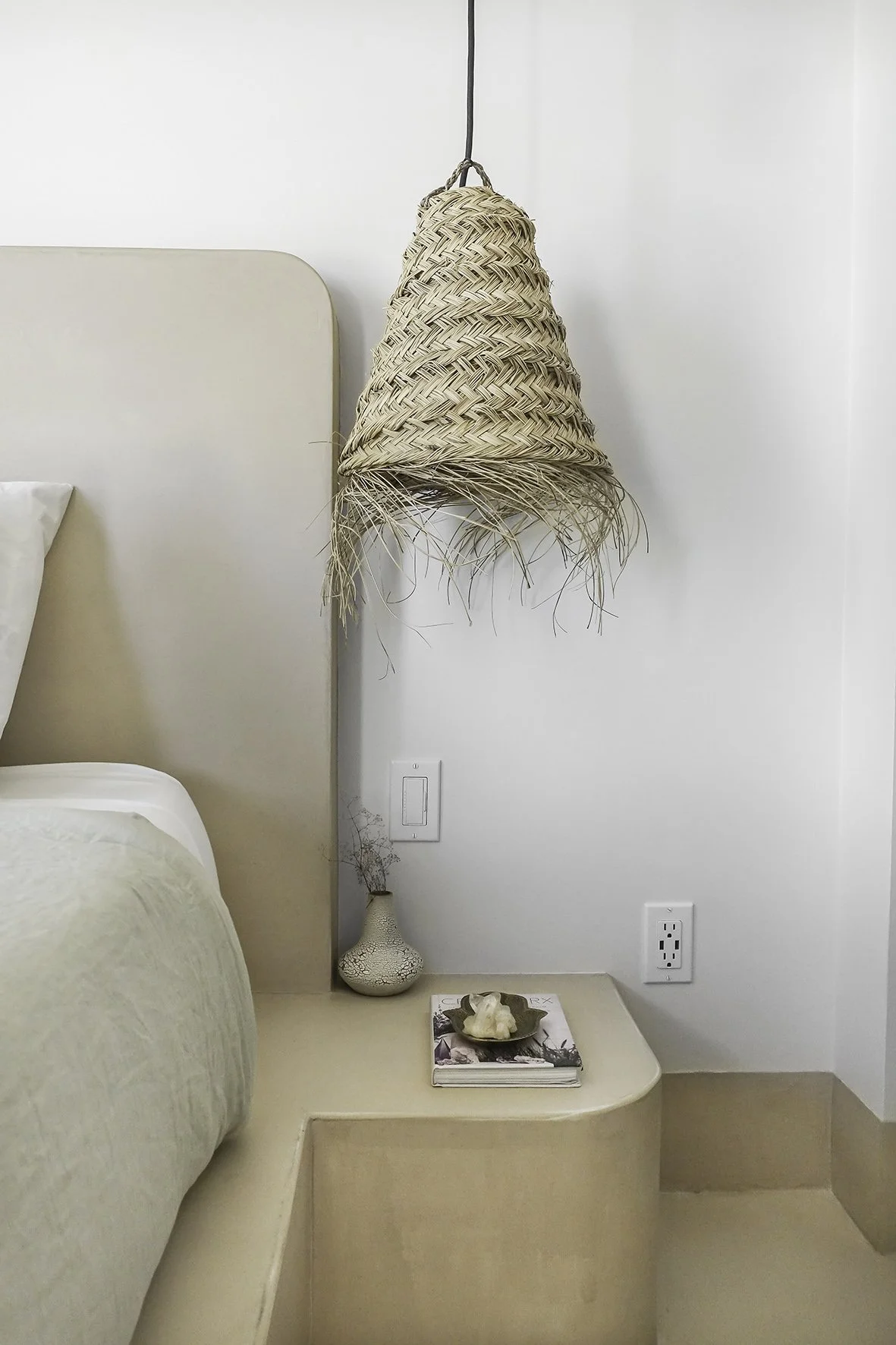 a custom poured-plaster bed frame with side tables, all as one piece with a woven hanging pendant, pot and magazines in a pile